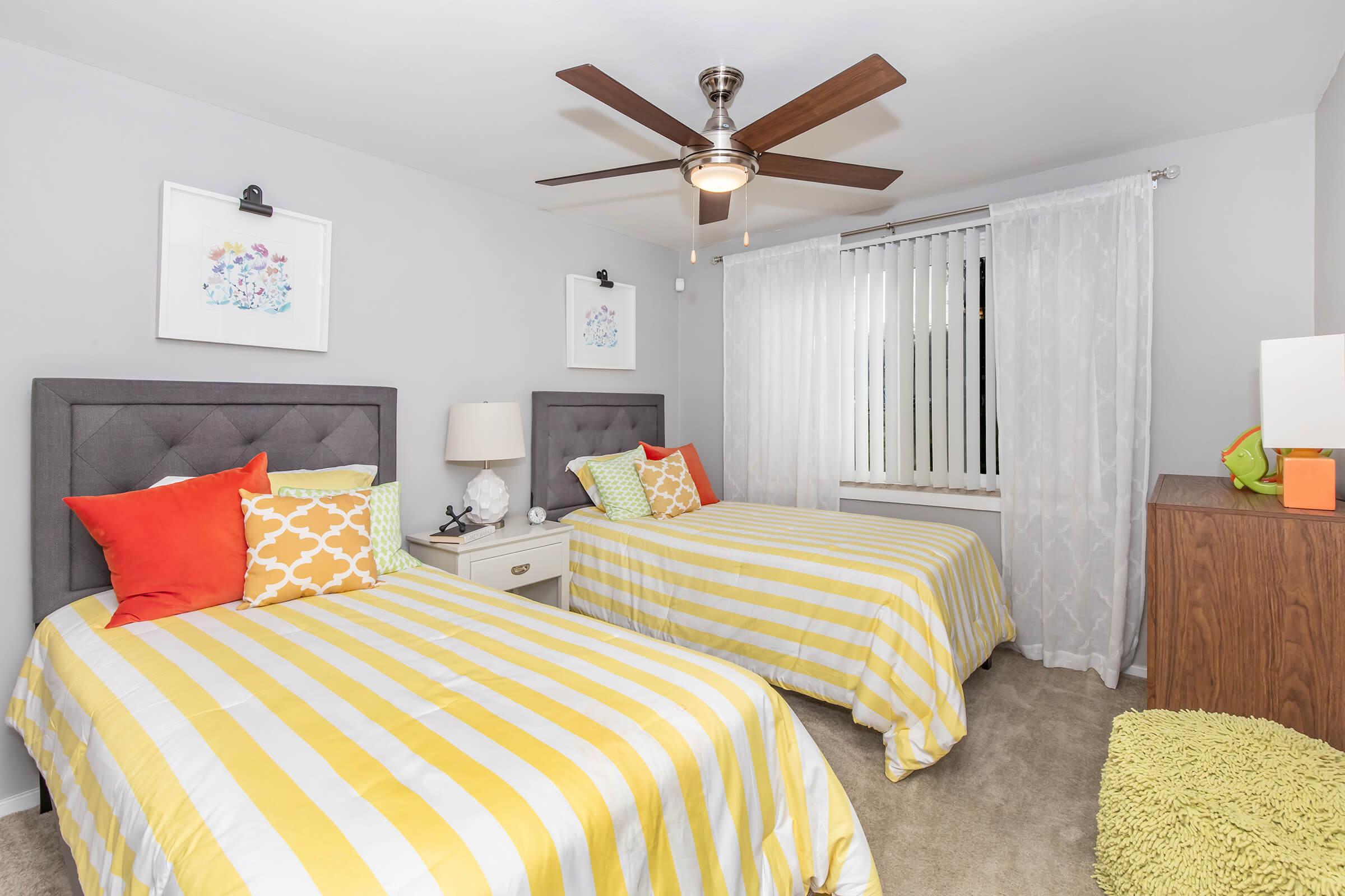 A bright and inviting bedroom featuring two twin beds with yellow and white striped bedspreads. Each bed has colorful pillows in shades of orange and green. There is a nightstand with a lamp between the beds and decorative wall art above them. A ceiling fan hangs overhead, and a window with sheer curtains lets in natural light.