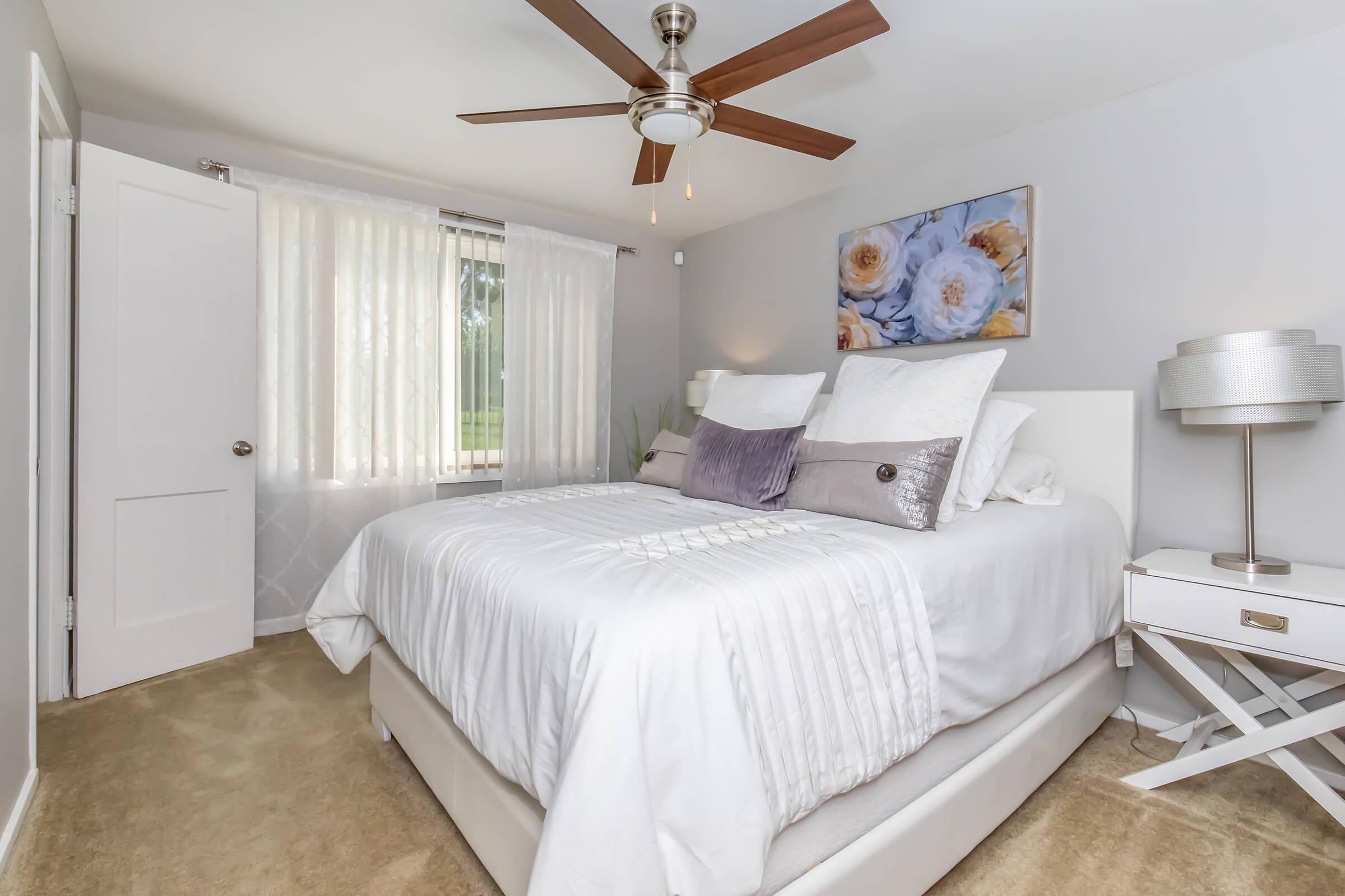 A cozy bedroom featuring a queen-size bed with white bedding, decorative pillows, and a light-colored headboard. A ceiling fan provides ventilation, while sheer white curtains allow natural light. A side table with a lamp complements the decor, and a wall art piece with floral designs adds a touch of elegance.
