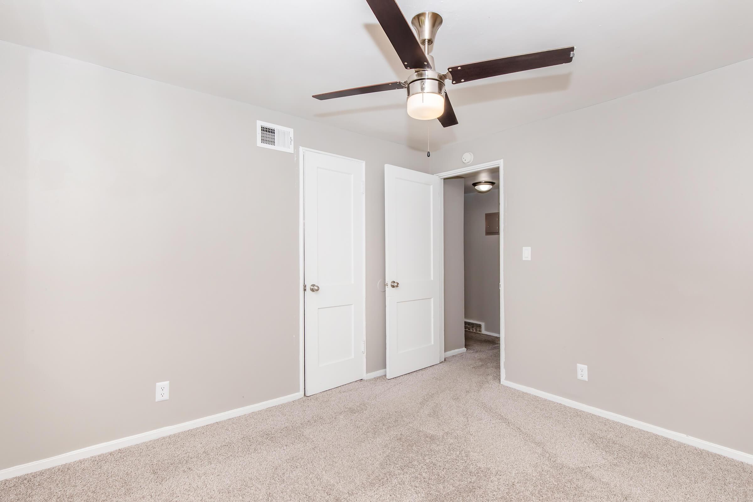 A light-colored room with beige carpet, featuring a ceiling fan with a light fixture. Two closed white doors are on the left side, and a painted wall and a light switch are visible. The room has a minimalistic design and is well-lit, creating an open, airy atmosphere.