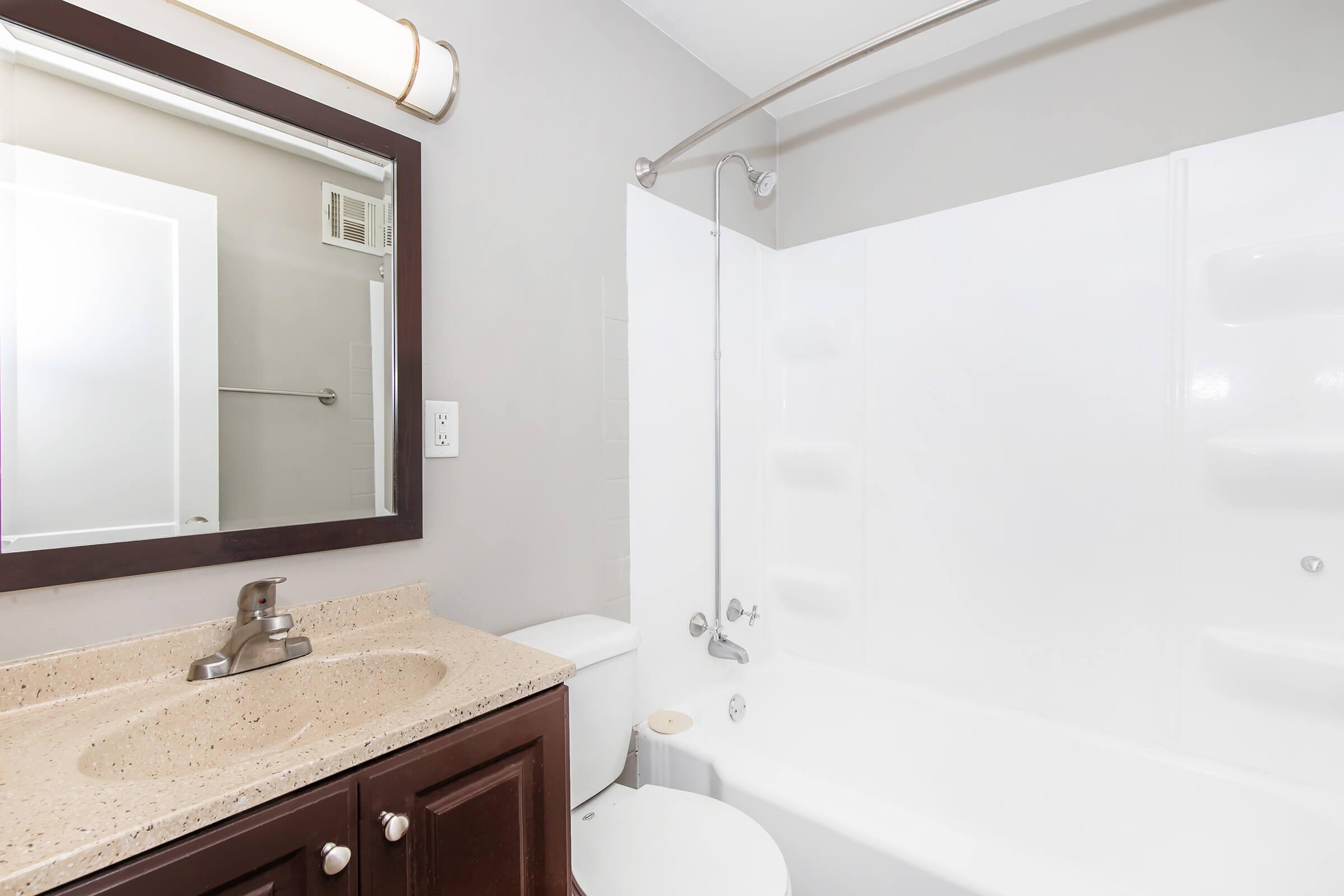 A clean bathroom featuring a white bathtub with a showerhead, a dark wood vanity with a sink, and a large mirror above. The walls are painted a light gray, and there's a small window letting in natural light. A towel rack is mounted to the wall opposite the shower.