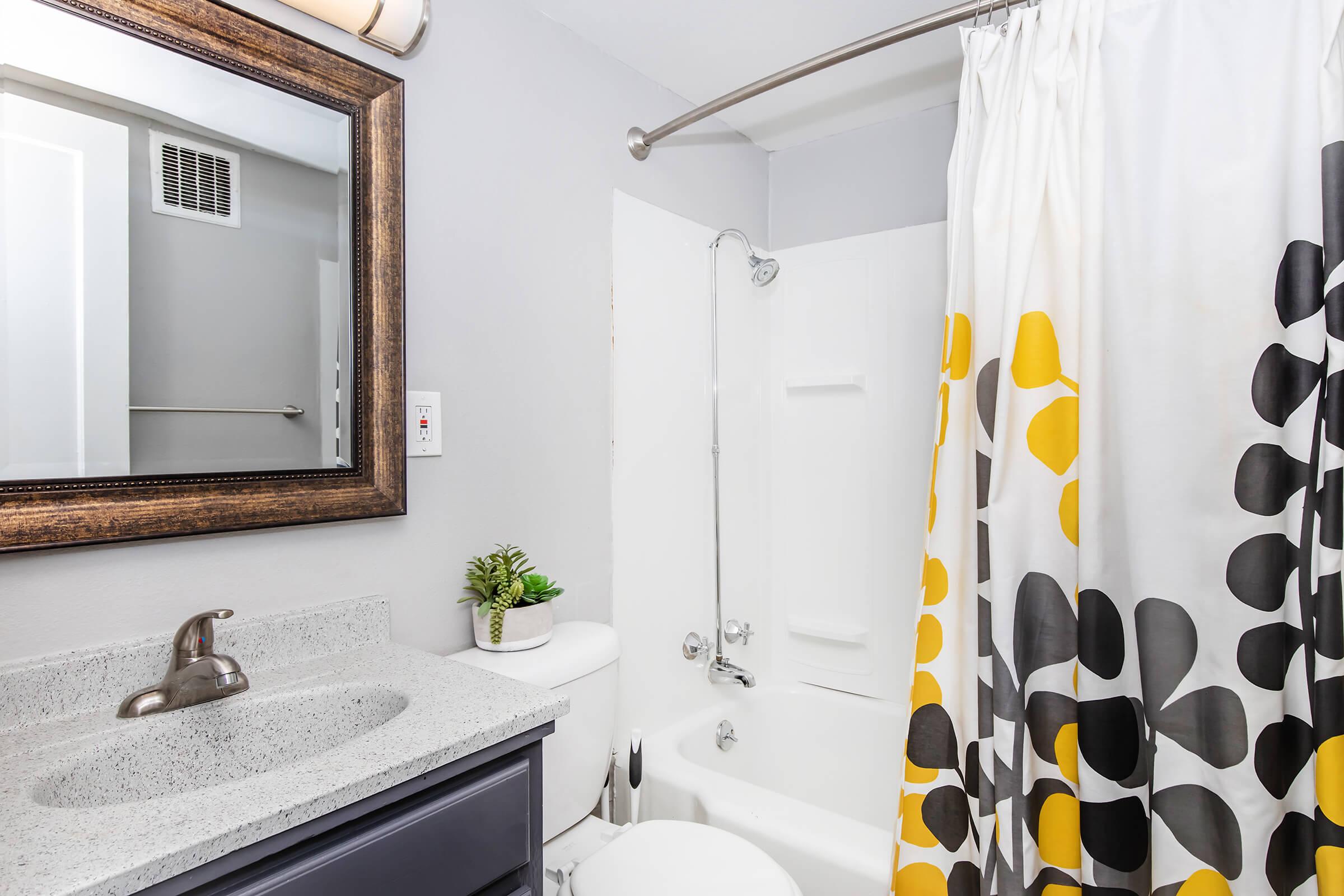 A modern bathroom featuring a bathtub with a shower, a patterned shower curtain in yellow and black, a sleek countertop with a faucet, a decorative plant, and a large mirror framed in dark wood. The walls are painted light gray, enhancing the contemporary feel of the space.
