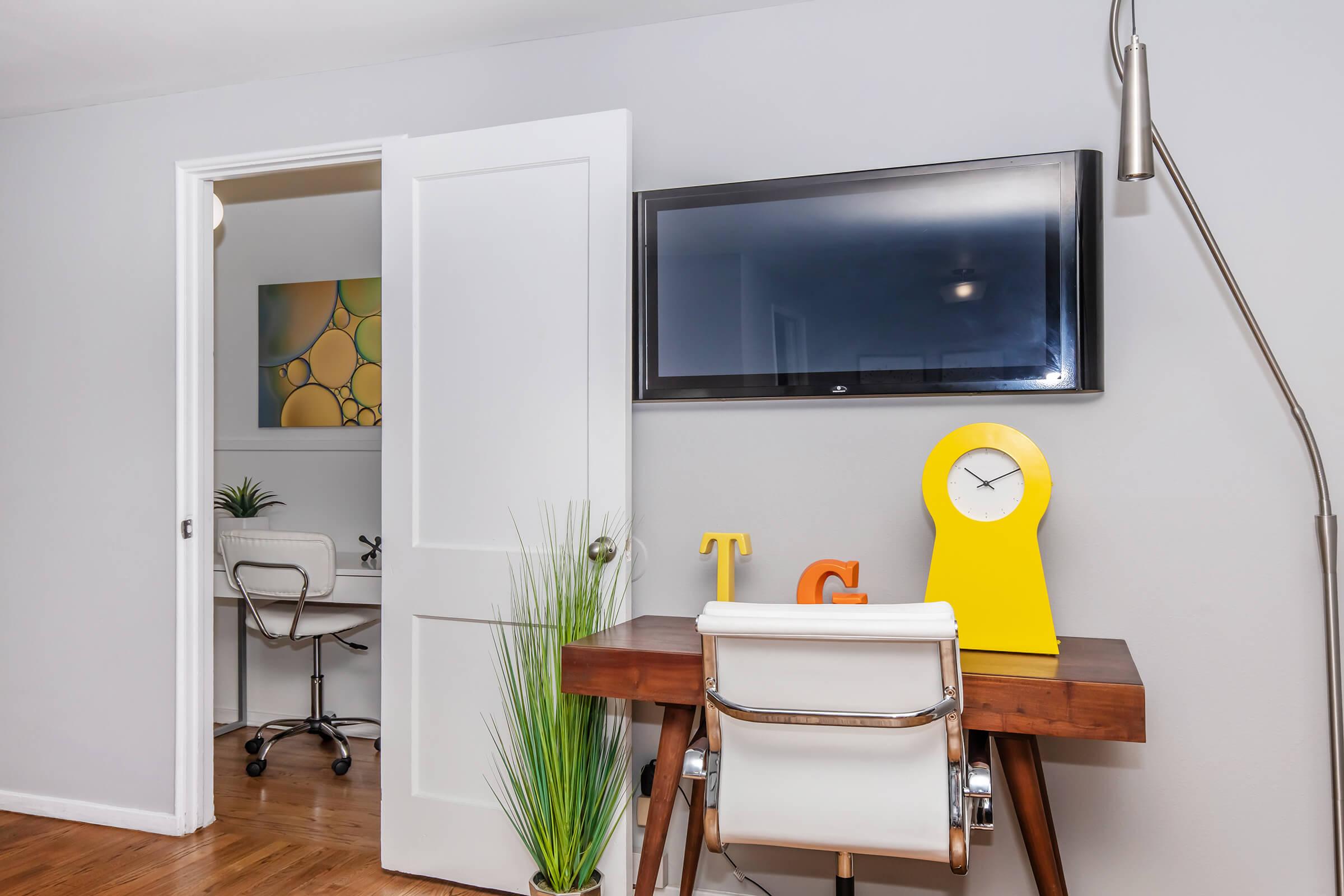 A modern home office setup featuring a sleek wooden desk with a yellow clock and decorative letters. A comfortable white chair is positioned at the desk. A wall-mounted TV is on the wall above the desk. In the background, a partially open door leads to another room with an additional desk and plants in both spaces.