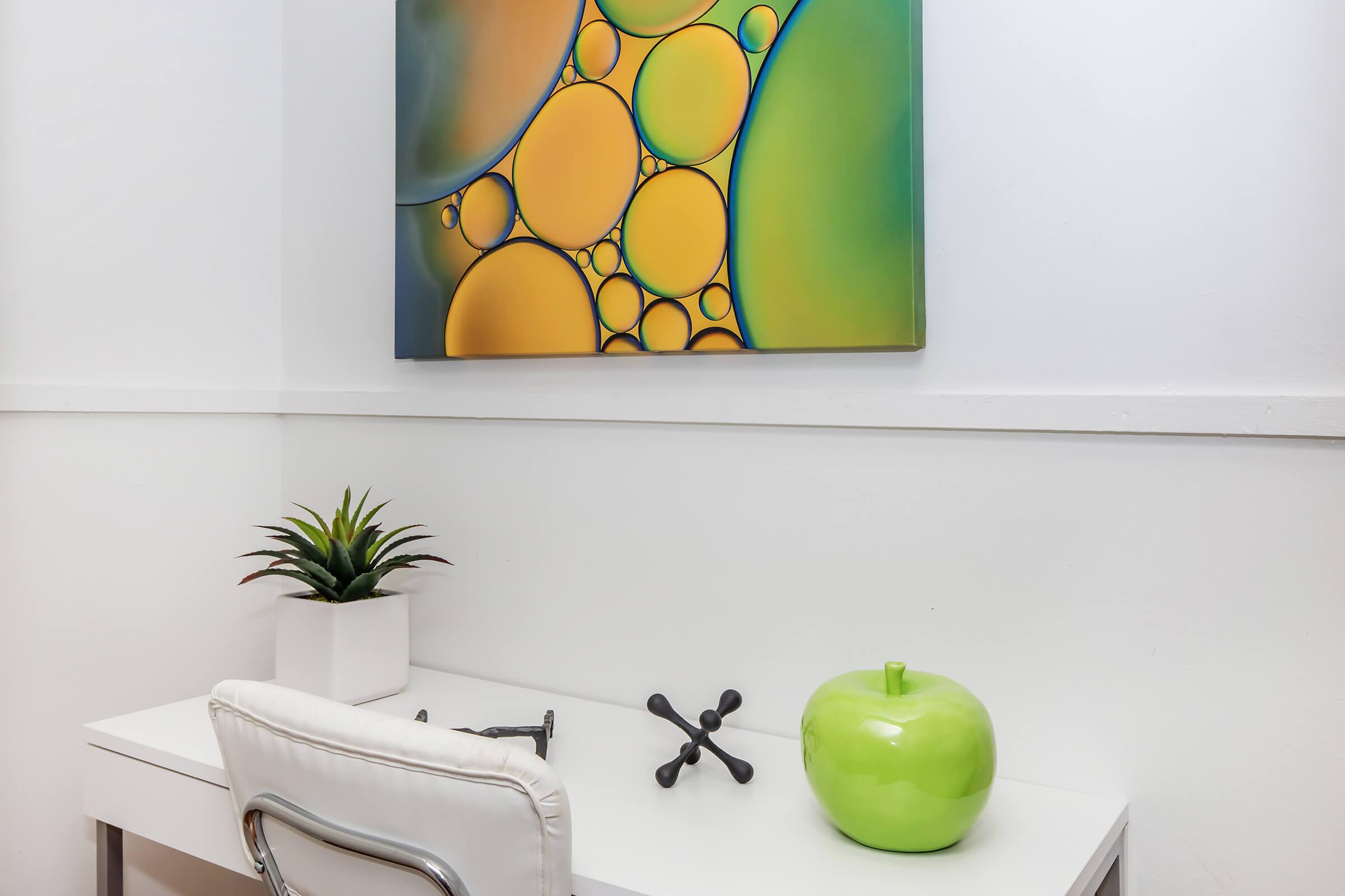 A minimalist workspace featuring a white desk with a sleek chair, an abstract painting with yellow and green circles on the wall above, a small potted plant in a white container, and a decorative green apple sculpture.