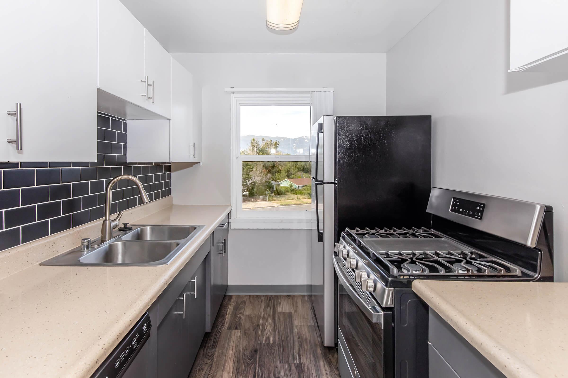 Modern kitchen featuring a sleek design with gray cabinets, a stainless steel gas range, and a double sink. The backsplash is black tile, and the countertops are light-colored. A large window offers a view of greenery outside, contributing to natural light in the space.