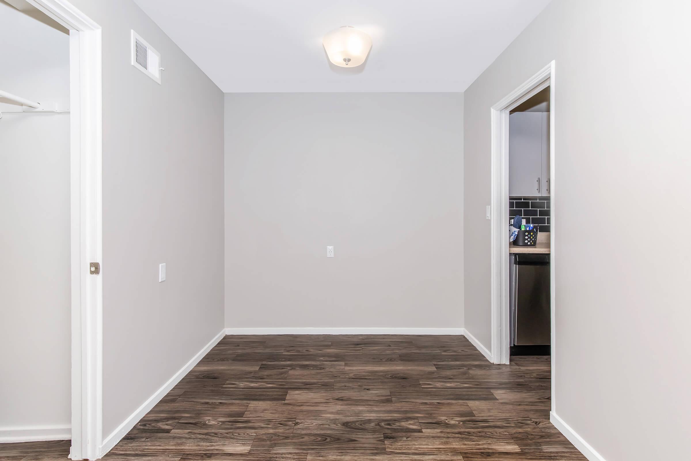 A blank interior room with light gray walls and dark wood flooring. A single light fixture hangs from the ceiling. Two doors are visible: one leads to a closet, and the other opens to a kitchen area. The space appears clean and uncluttered, suitable for various uses.