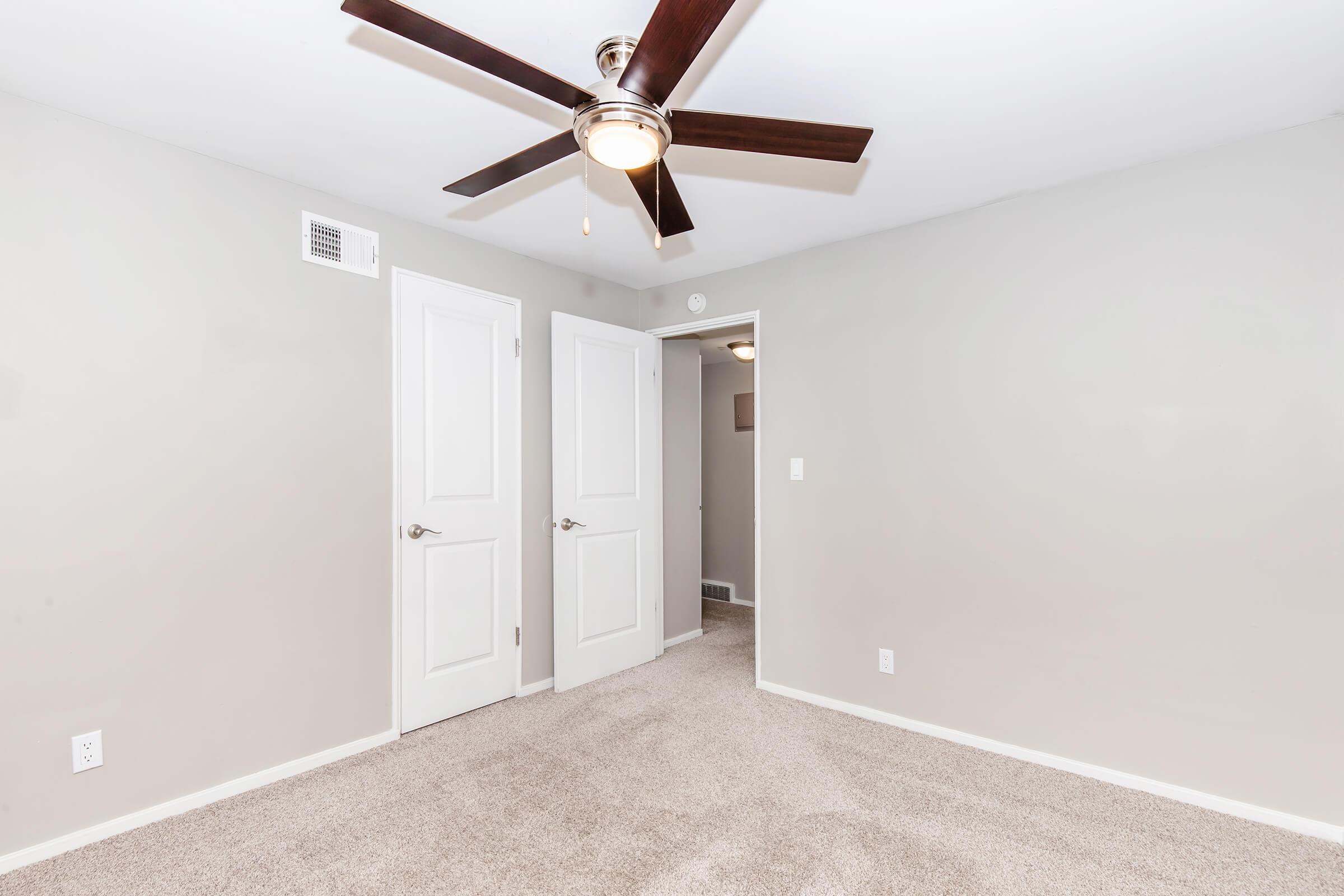 A neutral-toned room with beige carpet and light gray walls. It features a ceiling fan with five blades and two white doors leading to other spaces. The lighting is bright and the space appears empty and well-maintained, suggesting potential for various uses or decoration.