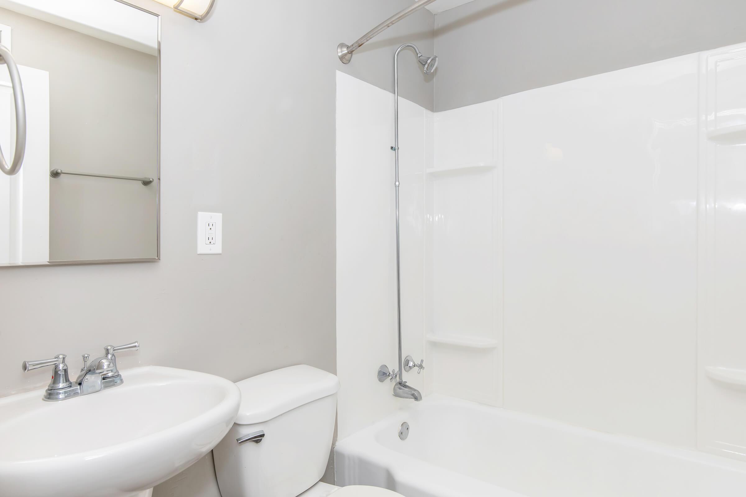 A clean and modern bathroom featuring a white bathtub with a shower, a sink with a chrome faucet, and a toilet. The walls are painted gray, and there's a mirror above the sink. The overall design is simple and functional, with ample lighting.