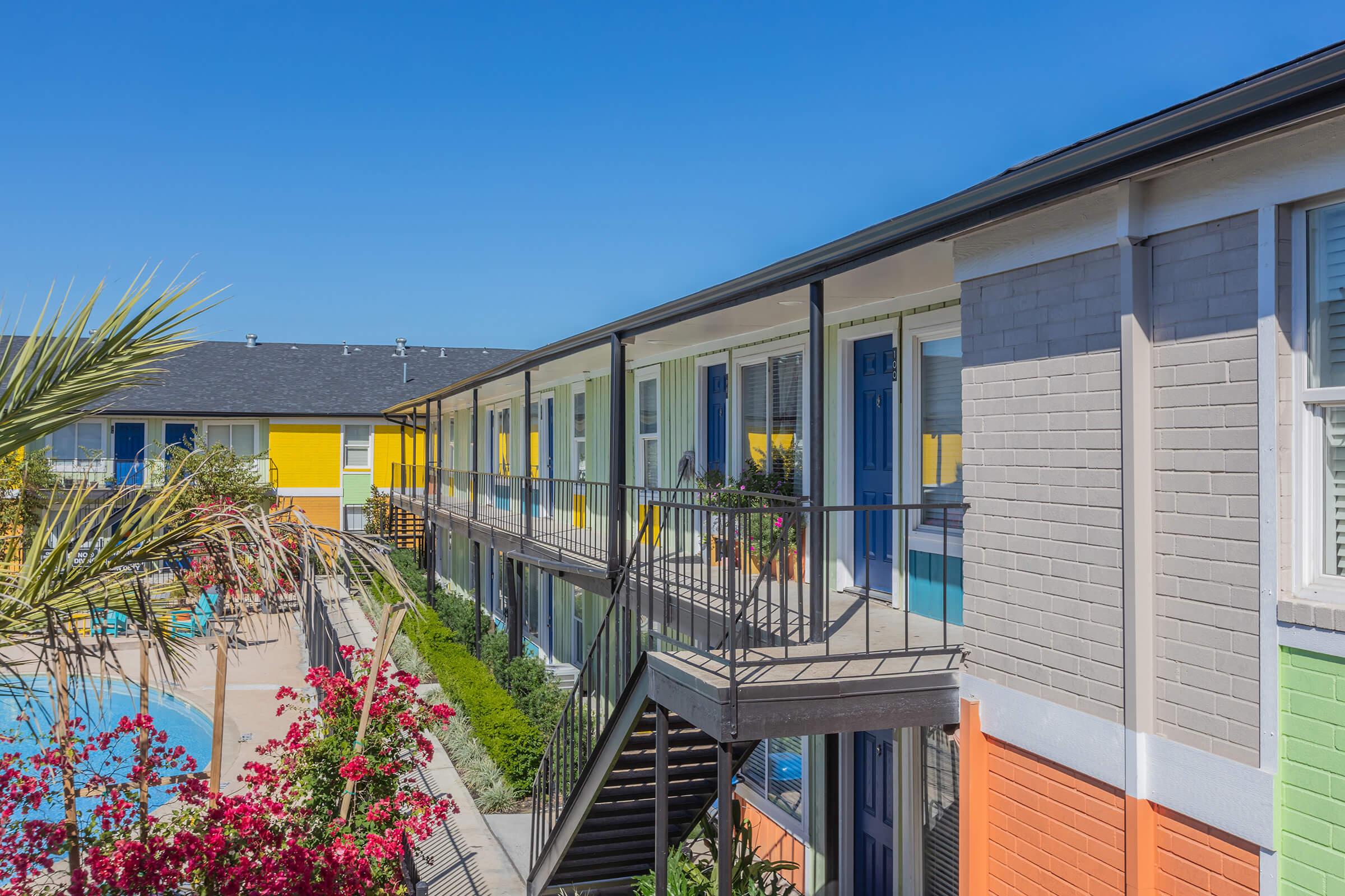 Brightly colored motel-style buildings line a pathway, with palm trees and vibrant pink flowers in the foreground. A swimming pool is visible nearby, and the clear blue sky adds to the cheerful atmosphere. The architecture features a mix of colorful facades and stairways leading to upper levels.