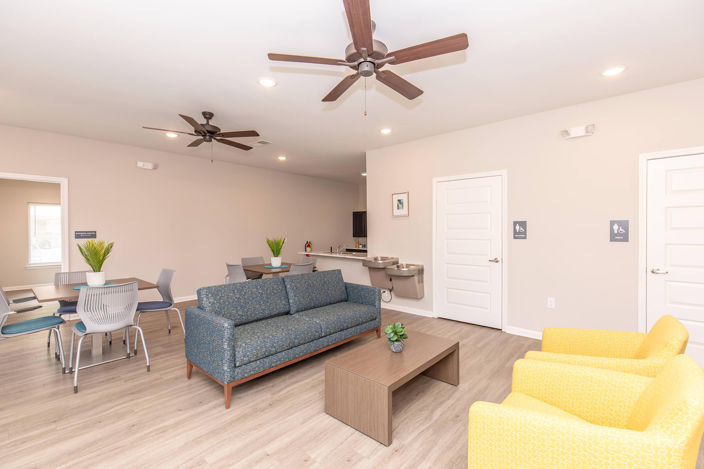 A spacious communal room featuring a blue sofa, two yellow armchairs, and a wooden coffee table. In the background, there are dining tables with chairs, a kitchenette area, and ceiling fans. The walls are painted in a light color, creating a bright and inviting atmosphere.