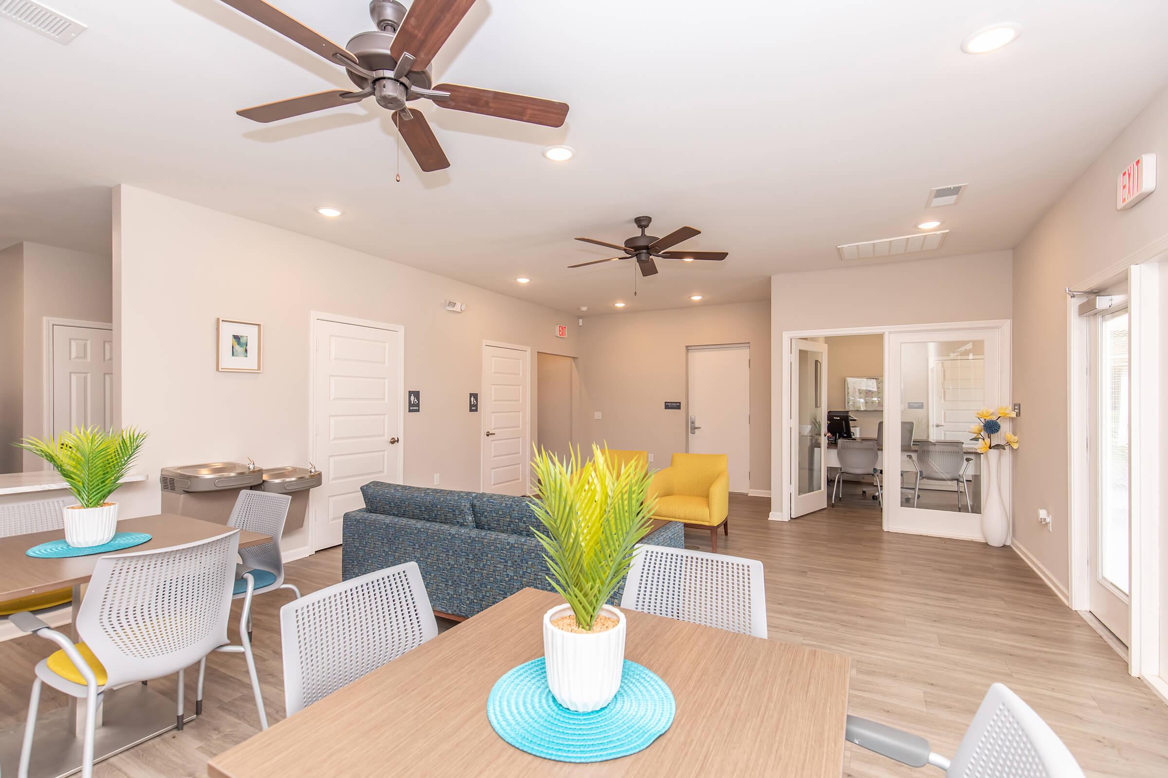 A modern lounge area featuring a large table with chairs, a blue sofa, and a bright yellow accent chair. Two ceiling fans are visible, and there are potted plants on the tables. The room has light-colored walls and hardwood flooring, creating a welcoming atmosphere. Glass partitions lead to another room.