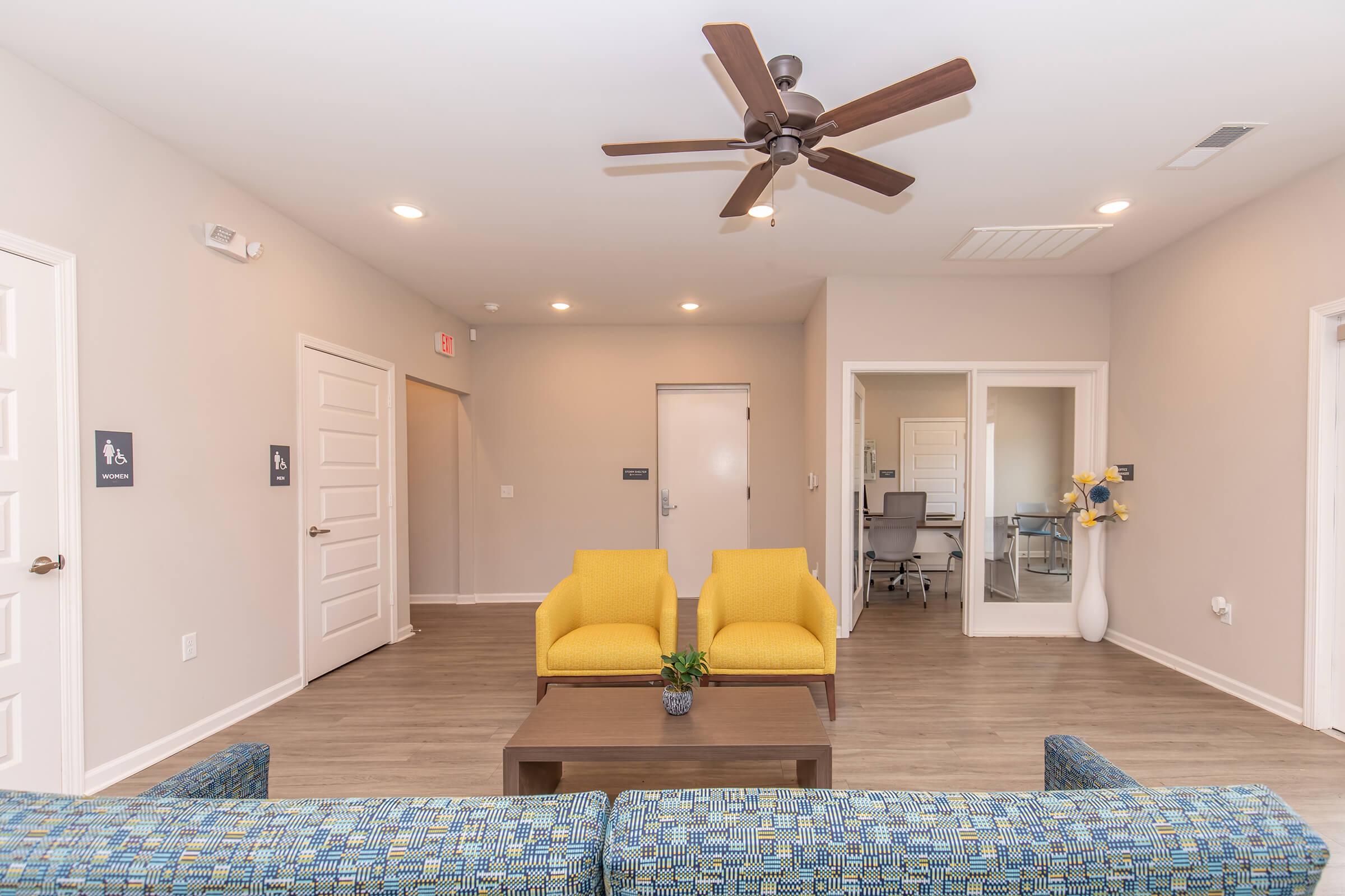 A spacious, modern waiting area featuring a blue patterned sofa and two yellow chairs around a low wooden table. A ceiling fan provides ventilation. The walls are painted light gray, and there are signs on the walls indicating restrooms. A door leads to an office area in the background.