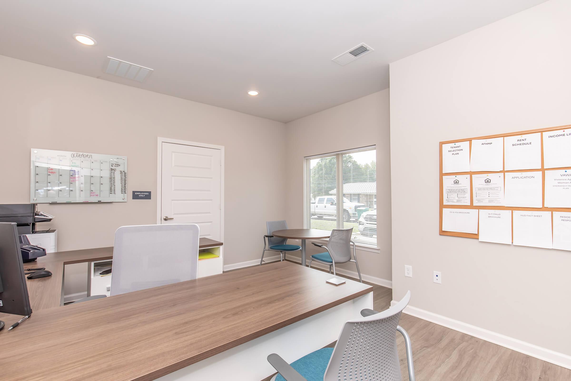 A modern office interior featuring a wooden desk, gray chairs, and a bulletin board with documents. There is a glass window allowing natural light, and a whiteboard is visible on the wall, adding a professional touch to the space.