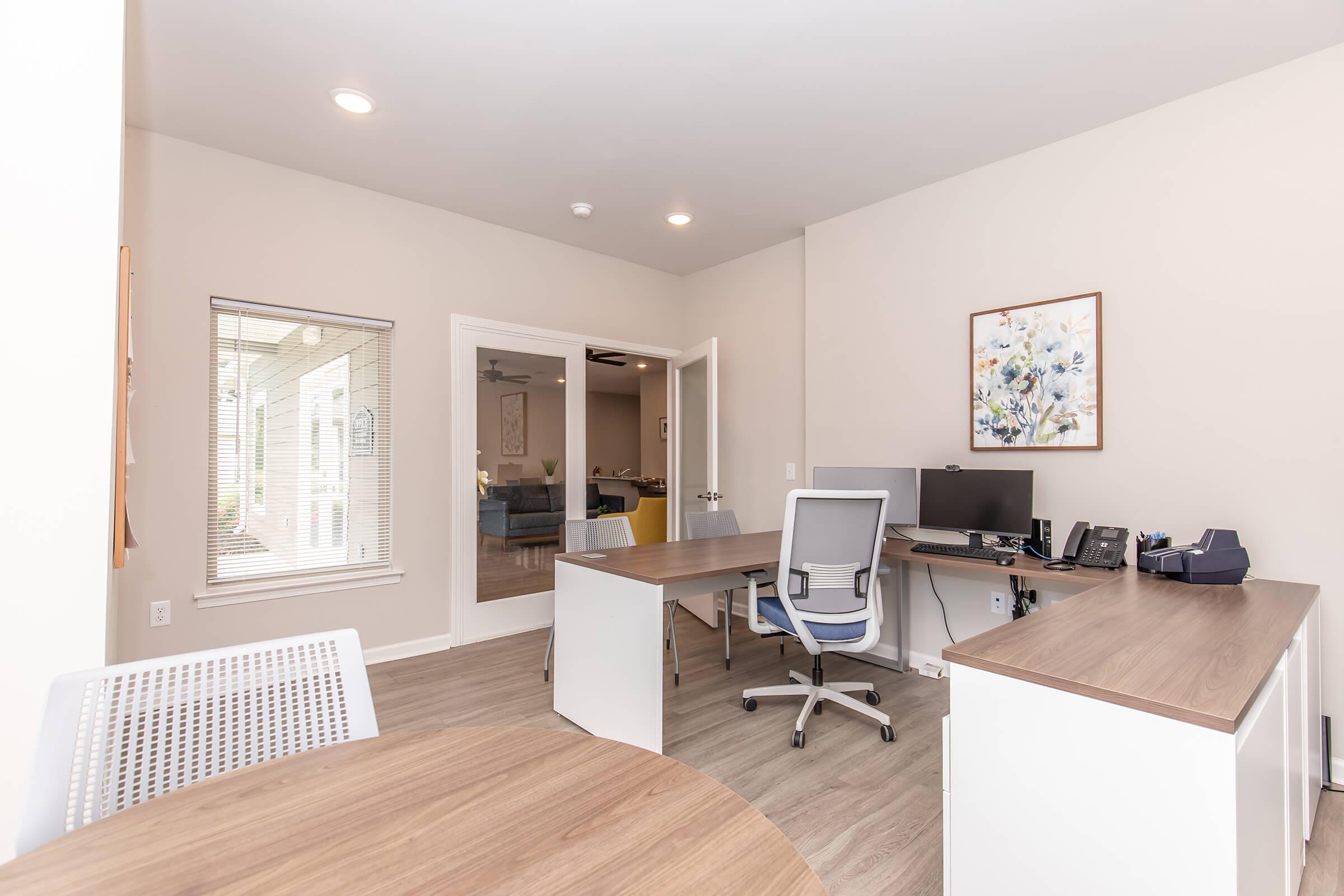 A modern office space featuring a wooden desk with a computer setup, an office chair, and a round table. Natural light streams in through a window, and there's a door leading to a separate room. The decor is simple and minimalistic, with a floral artwork on the wall.