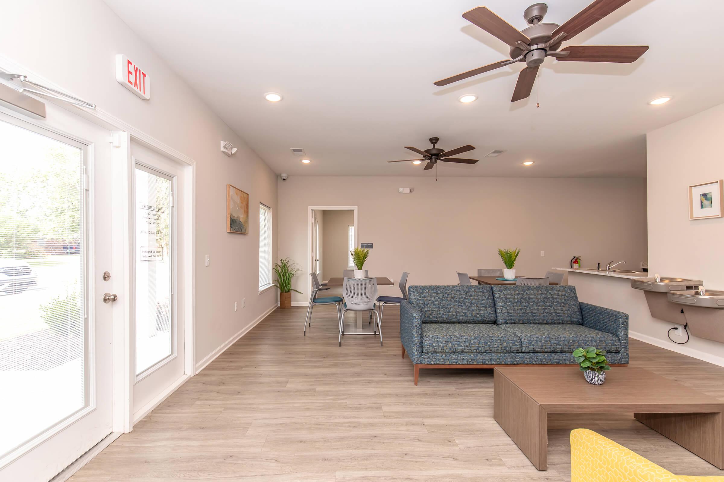 A spacious, modern living area featuring a blue sofa, a coffee table, and a dining table with chairs. The room has ceiling fans and large windows allowing natural light. In the background, there's a kitchenette area with a bar counter. Potted plants are placed around for added decor.