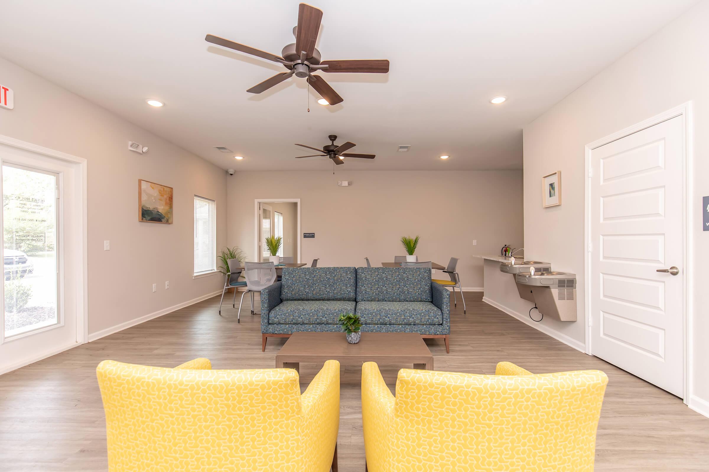 A modern lounge area featuring a blue couch, two yellow chairs, a coffee table, and a dining area with several chairs and tables. The space is well-lit with ceiling fans and has plants for decoration, creating a welcoming atmosphere.