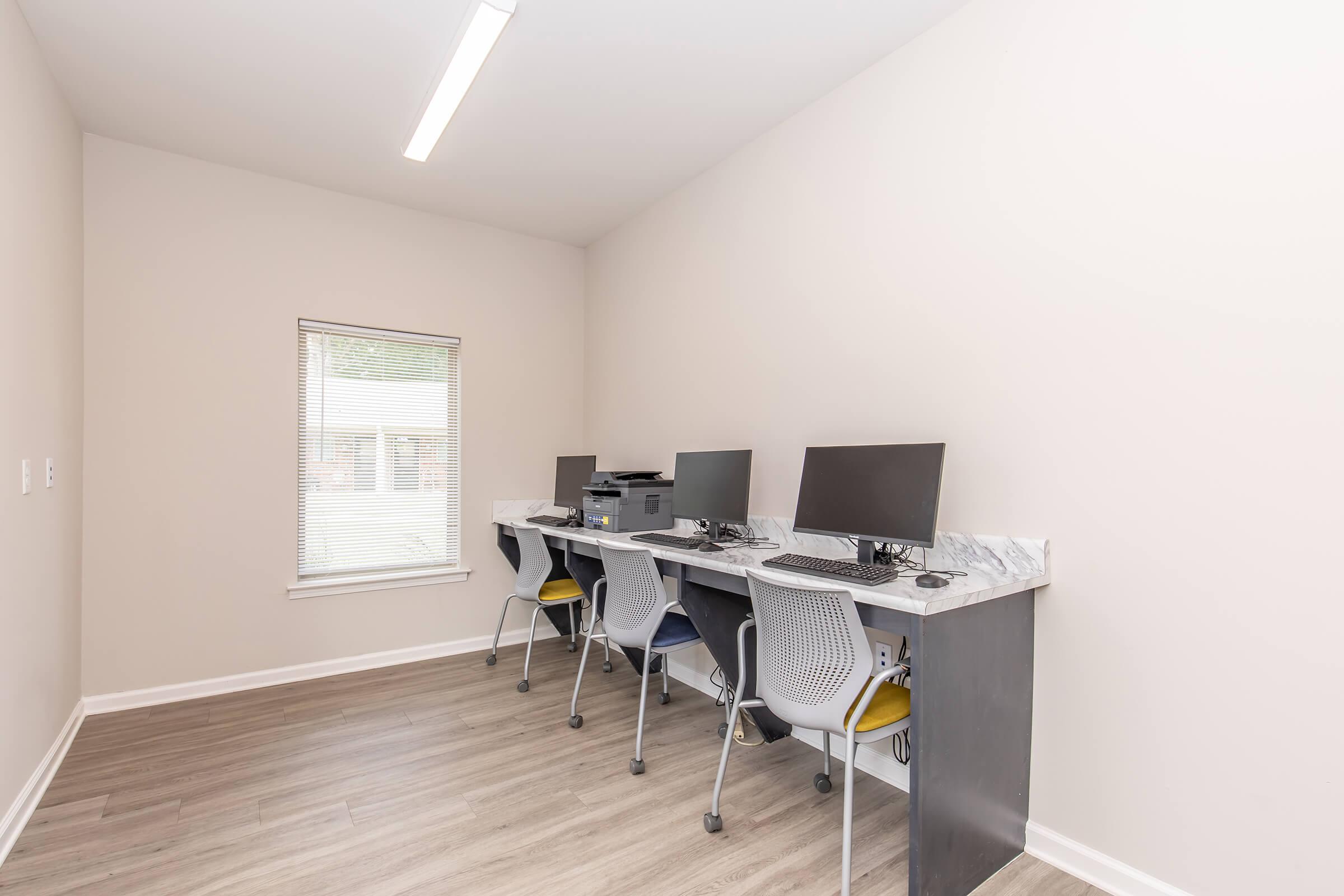 A minimalist office space featuring three computer workstations with monitors, a printer, and ergonomic chairs. The room has light-colored walls, wooden flooring, and a window allowing natural light to enter, creating a bright and functional environment for work or study.