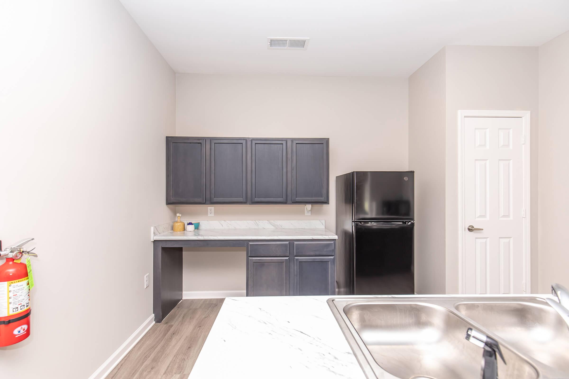 Interior view of a kitchen featuring dark cabinetry, a marble countertop, a black refrigerator, and a white door. A fire extinguisher is mounted on the wall to the left. The space has neutral-colored walls and a modern design, with a clean, minimalist aesthetic.