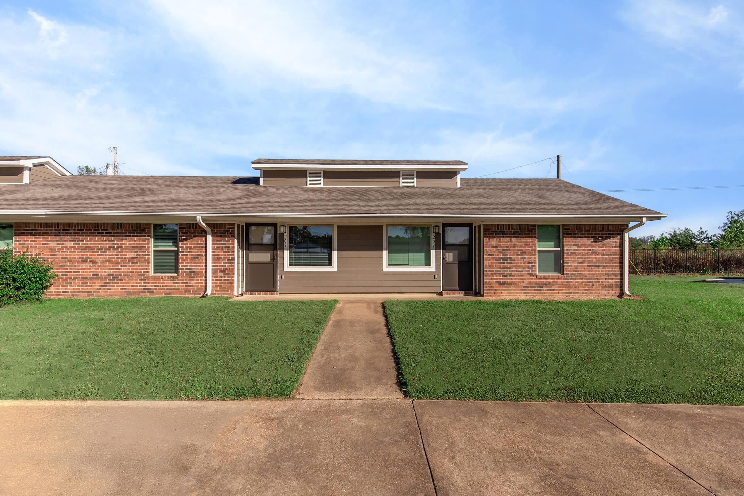 A single-story brick duplex with a tan and brown exterior. The building features two front doors, large windows, and a well-maintained lawn. A concrete walkway leads to the entrance, with a clear blue sky in the background.