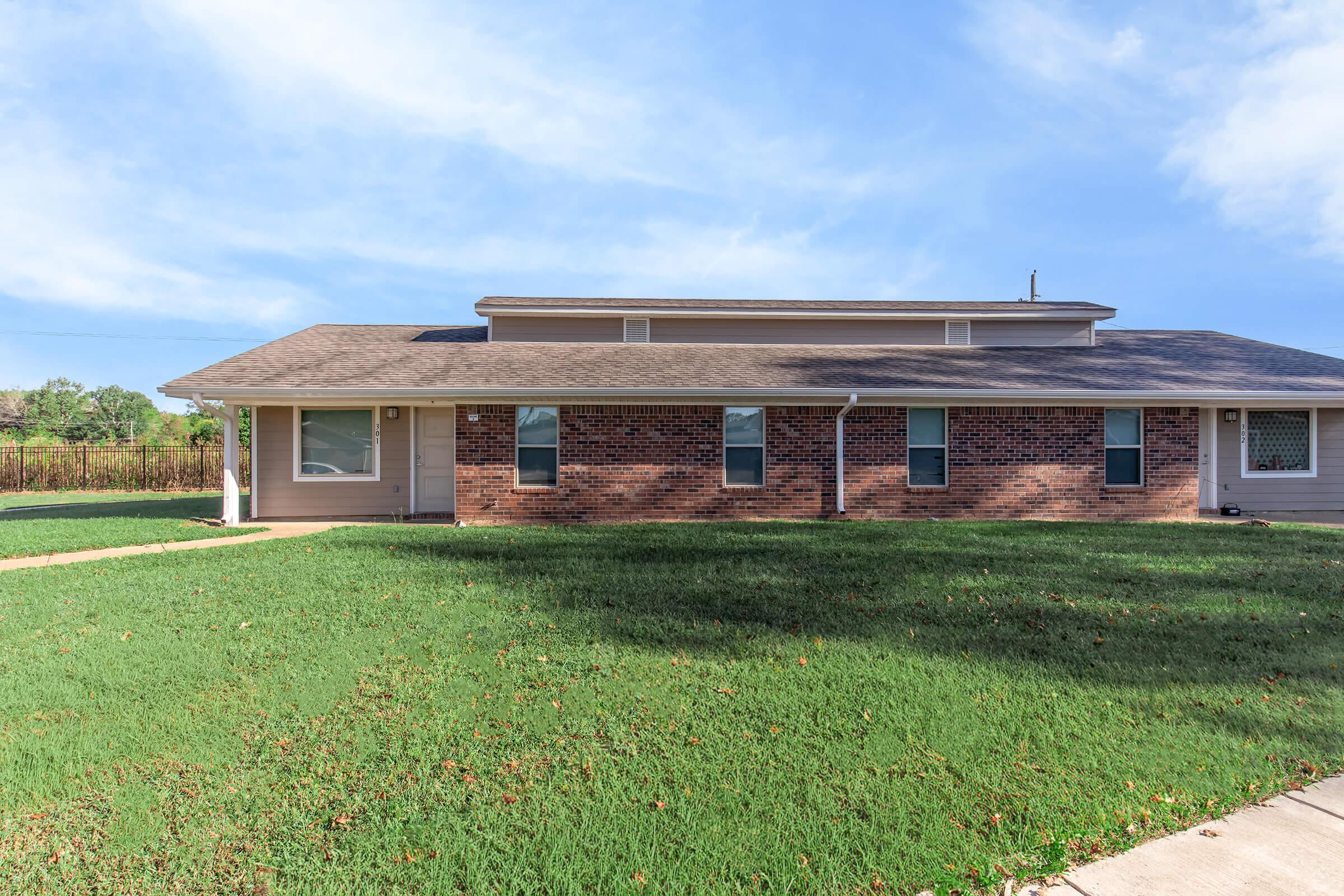 A single-story brick house with a pitched roof, featuring multiple windows and a well-maintained lawn. The driveway runs alongside the home, and there are a few trees in the background under a blue sky with wispy clouds. The property appears inviting and well-kept.
