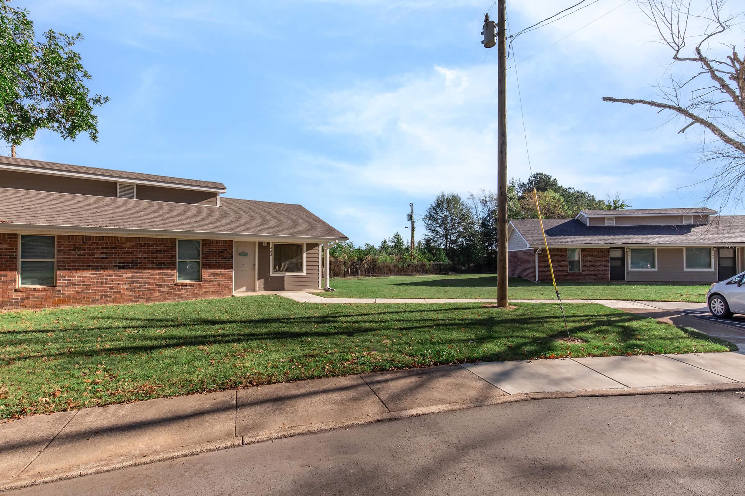 Two single-story residential buildings made of brick and wood, situated on a grassy lot. A sidewalk runs alongside the buildings, and a power pole is visible in the background. The scene is bright with clear blue skies and a few trees in the vicinity.