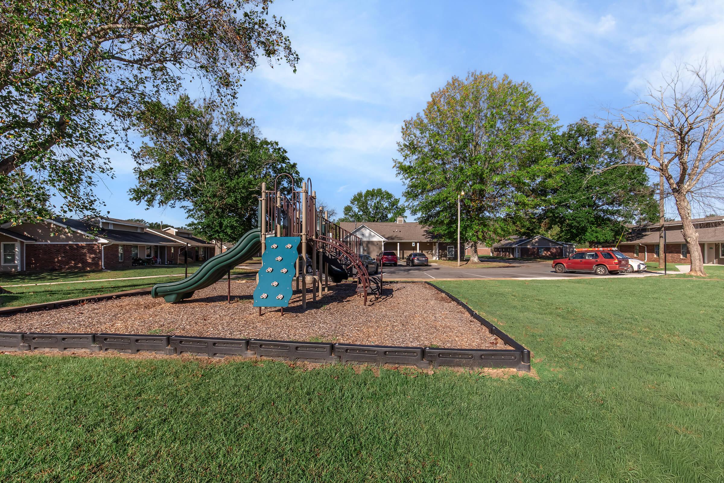 A playground with a slide and climbing structure surrounded by grassy areas and trees. Nearby residential homes are visible in the background, along with parked cars on a paved area. The scene is set on a clear day with blue skies.