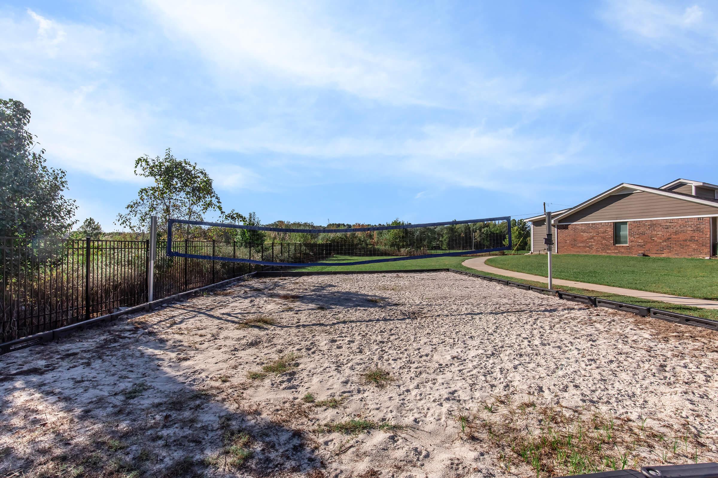 A sand volleyball court surrounded by a black fence, with a clear blue sky above. In the background, there are green trees and a pathway leading to a brick building, indicating a recreational area. The court is empty, suggesting it is ready for use.