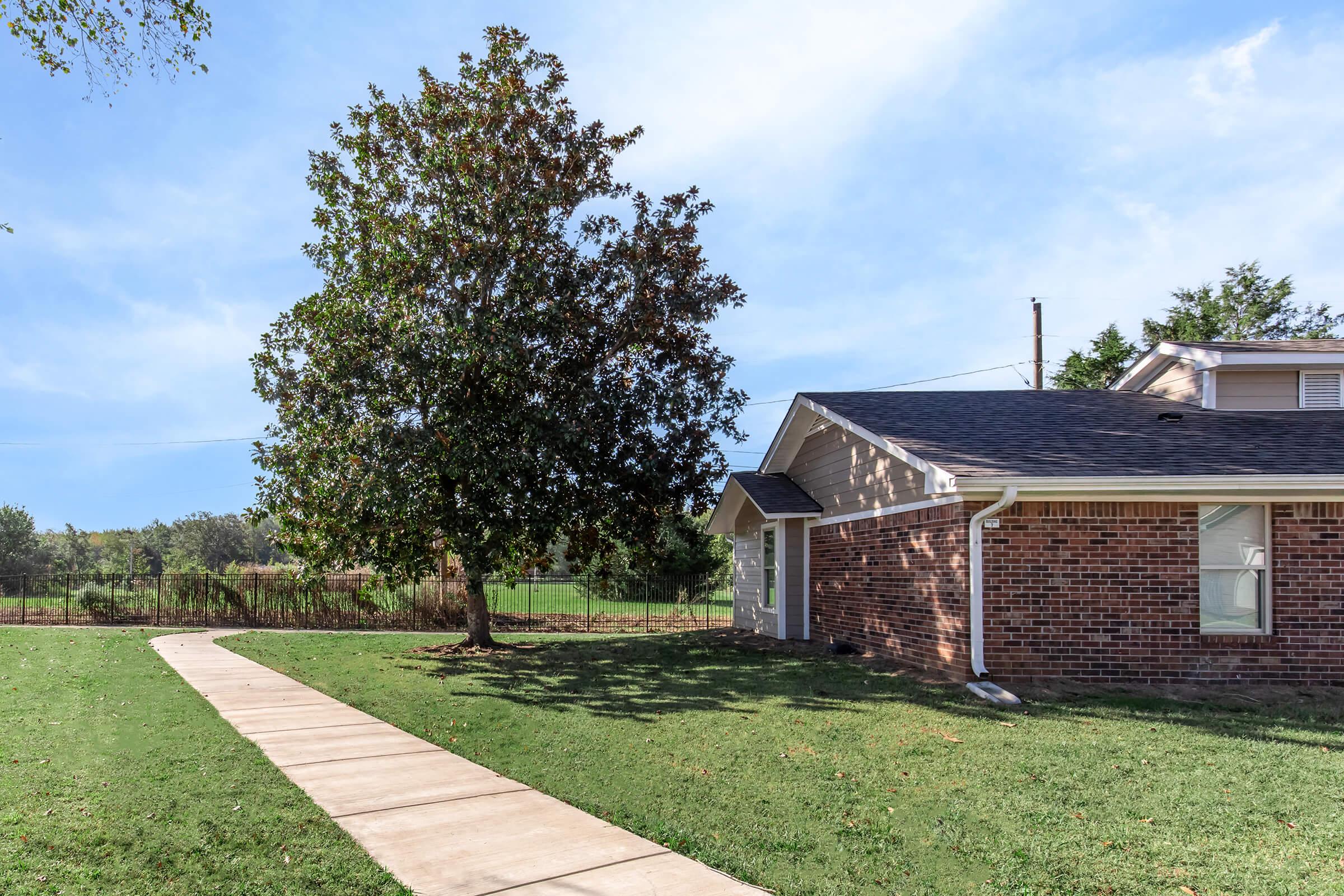 A brick house with a sloped roof stands beside a large green lawn, featuring a concrete path leading to the entrance. A large tree provides shade in the yard. In the background, a green field is partially visible under a clear blue sky with a few clouds.