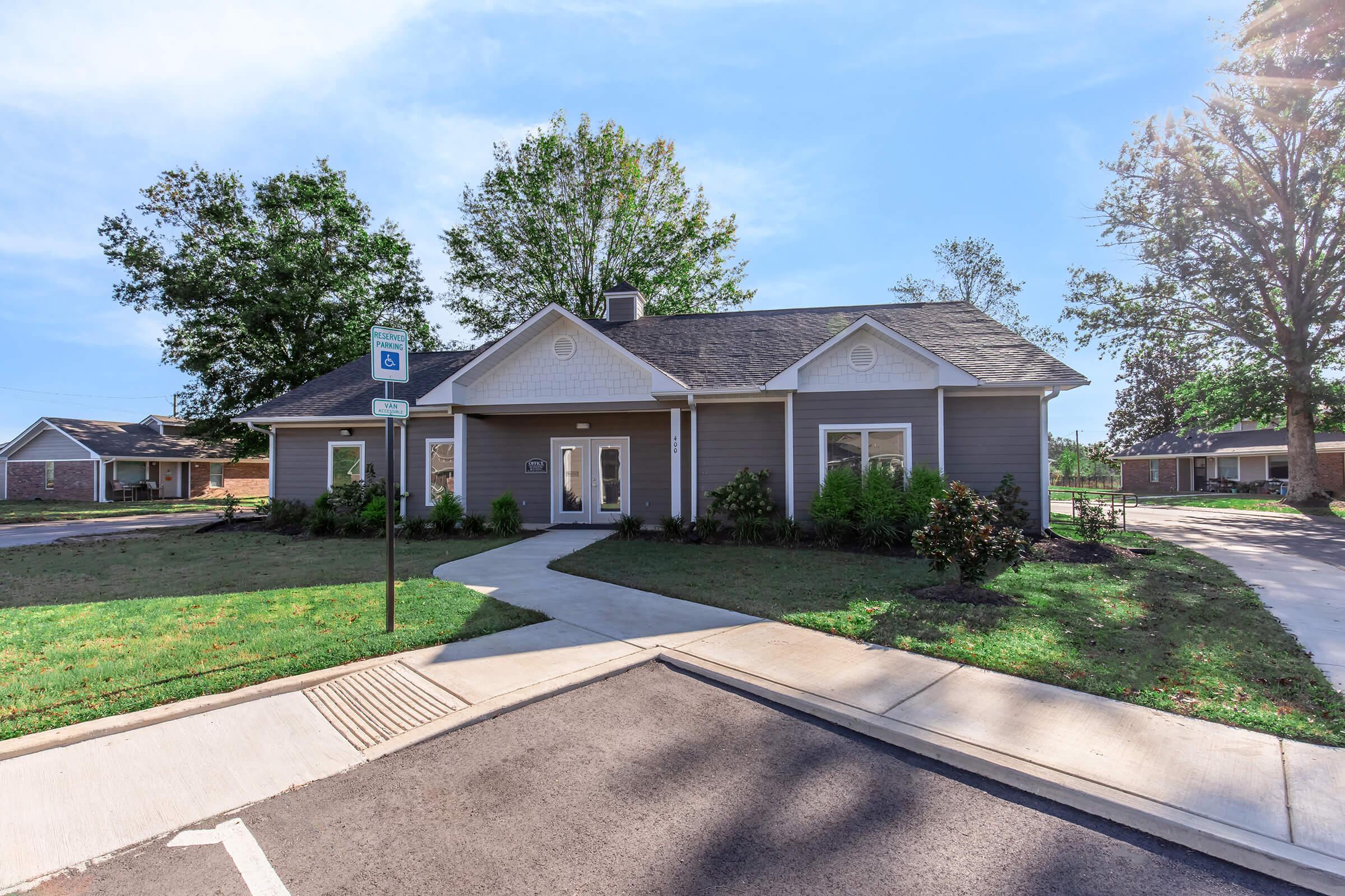 A single-story house with a gray exterior and a pitched roof, surrounded by green grass and trees. A paved pathway leads to the front entrance. There is a blue signpost nearby, and the area appears to be well-maintained and sunny, reflecting a residential neighborhood setting.