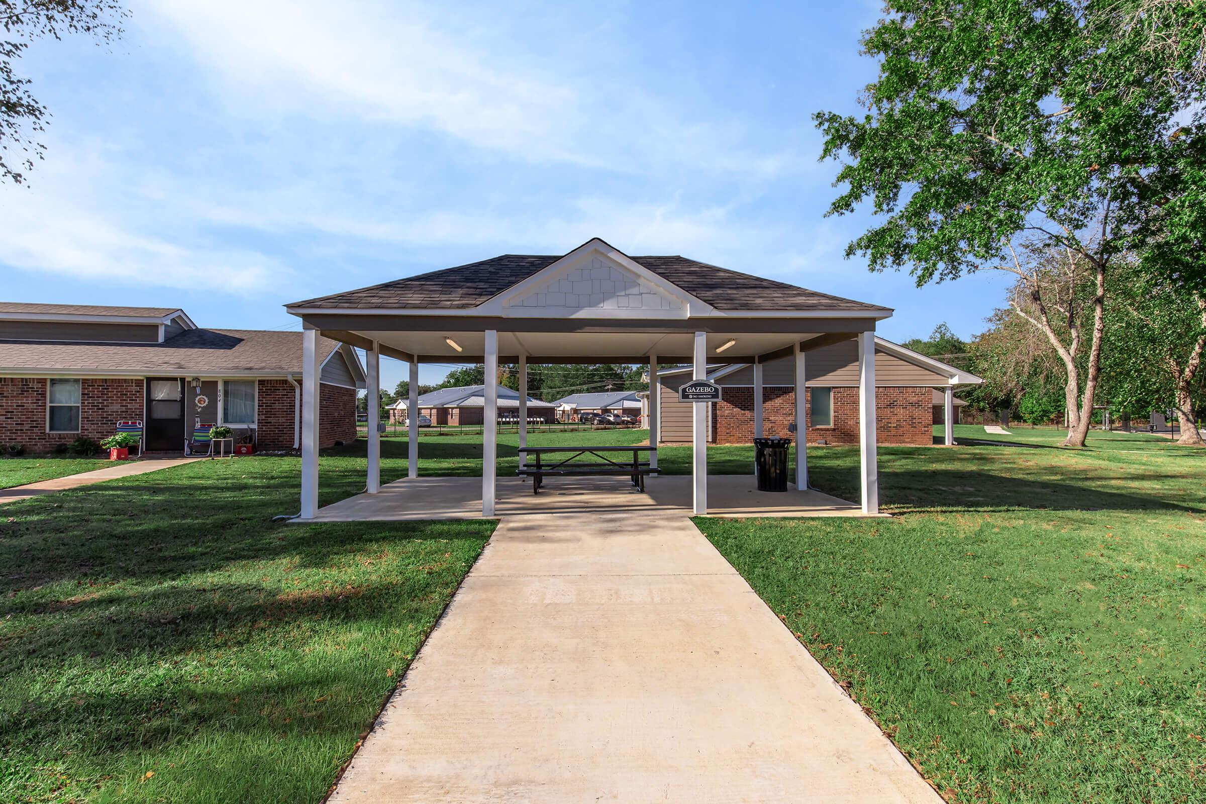 A paved pathway leads to a shaded pavilion with a picnic table, surrounded by neatly trimmed grass. In the background, there are brick buildings and trees. The sky is clear and blue, indicating a sunny day.