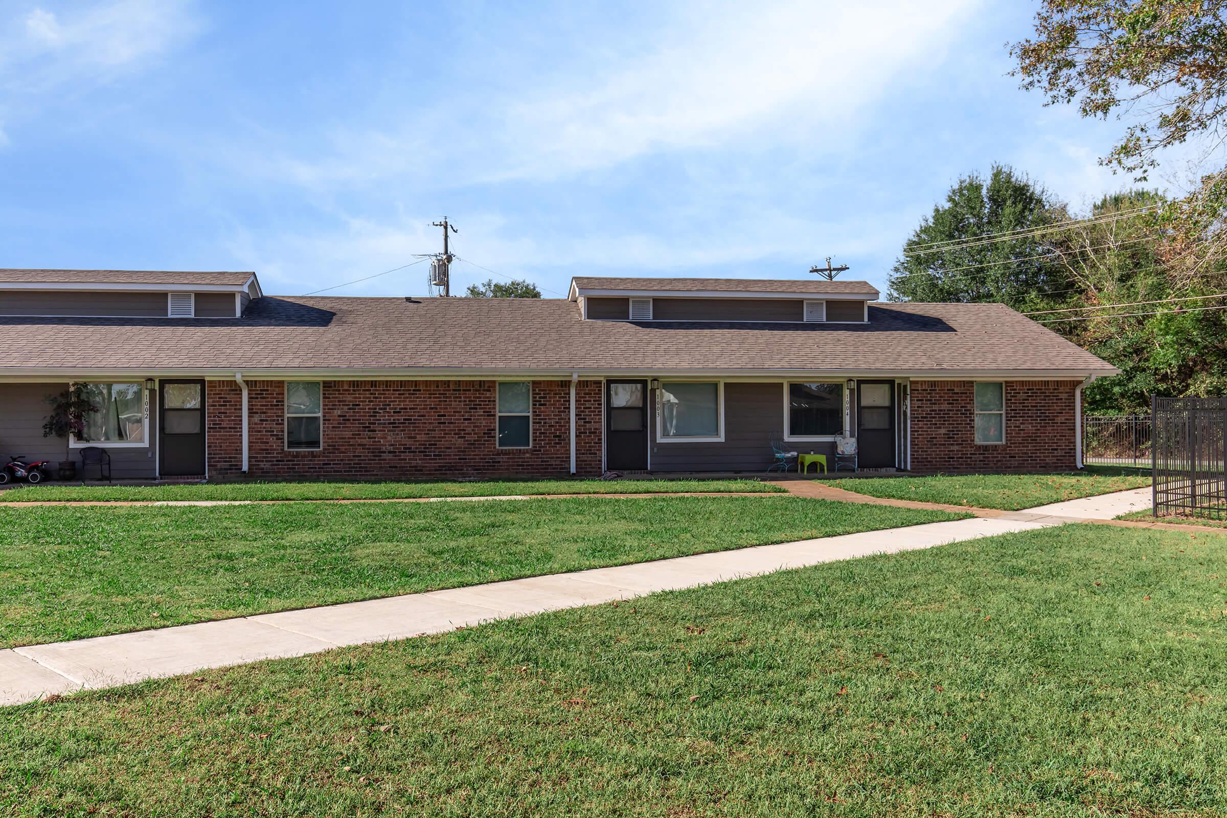A low-rise, brick apartment building with a sloped roof, featuring several front doors. A paved walkway runs through a green lawn in front of the building, with trees and power lines visible in the background under a clear blue sky. The scene conveys a quiet, suburban atmosphere.