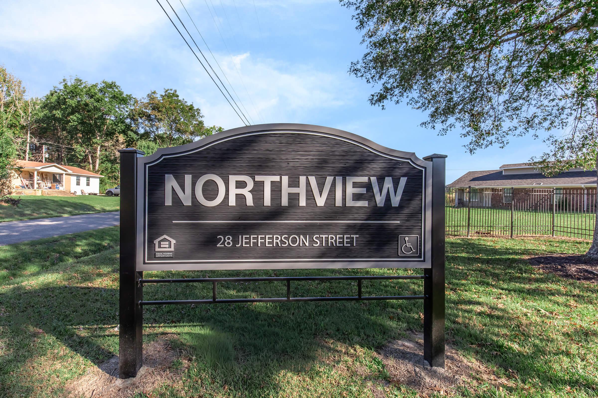 Sign displaying "NORTHVIEW" and "28 JEFFERSON STREET" against a black background. The sign is surrounded by green grass and trees, with houses visible in the background. There is a symbol indicating accessibility.
