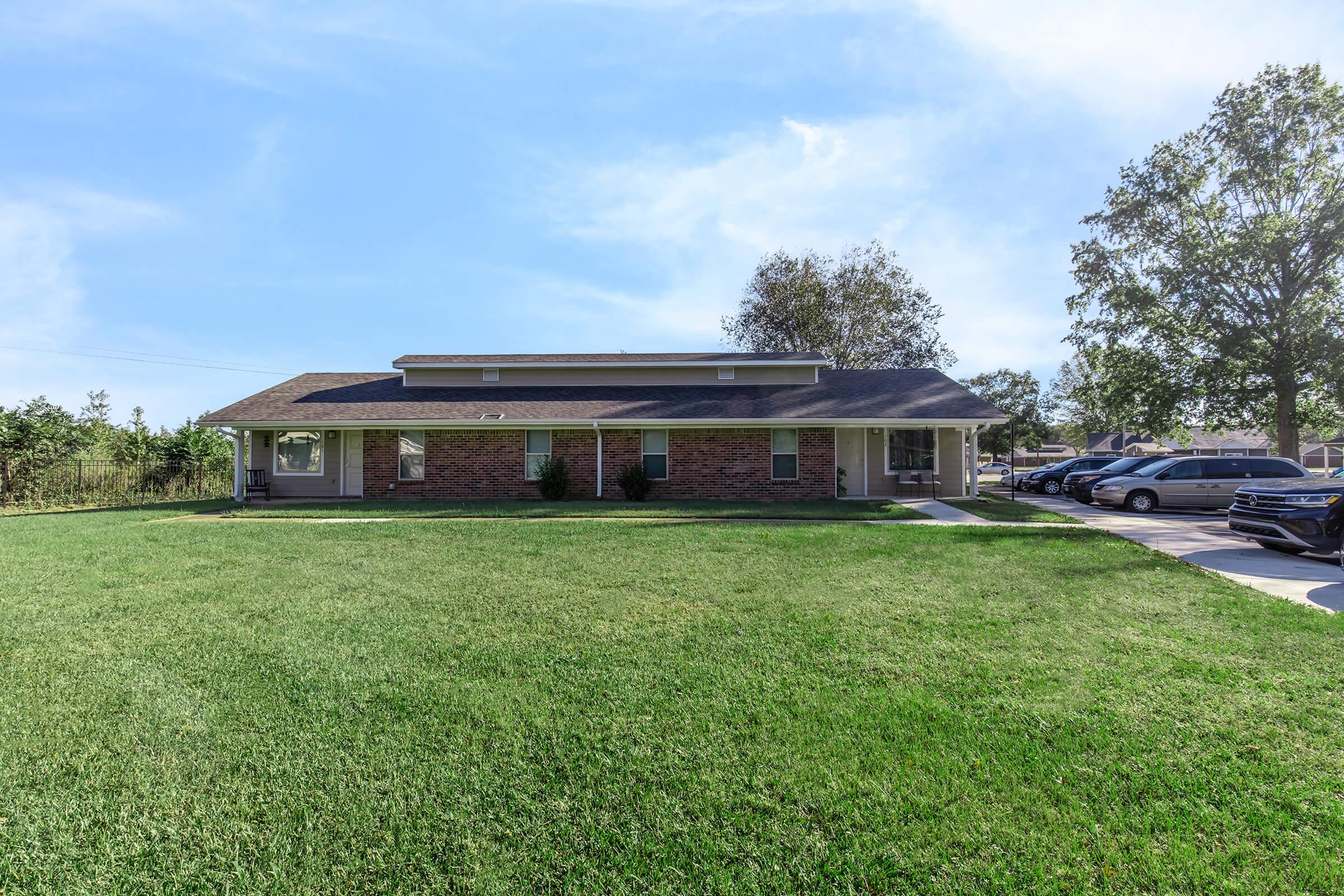 A single-story brick building with a flat roof, surrounded by a well-maintained lawn. The structure has a covered porch and several windows. Parked vehicles are visible in front, with trees and shrubs in the background under a clear blue sky.