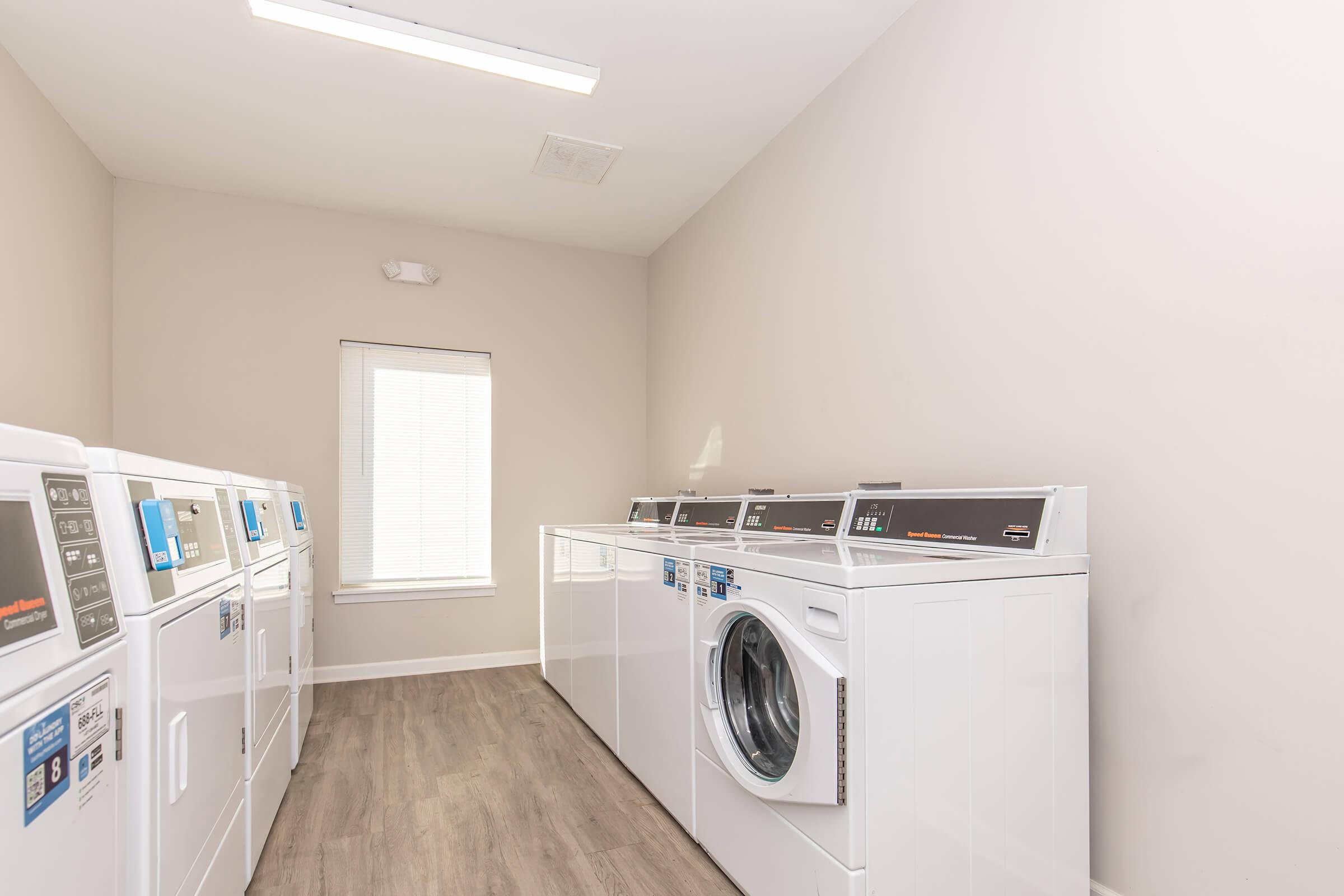 A brightly lit laundry room featuring several white coin-operated washing machines and dryers aligned along the wall. A large window provides natural light, and the room has light-colored walls and wooden flooring.