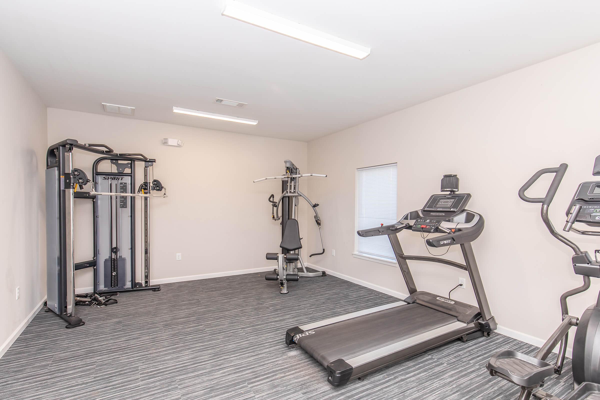 A modern home gym featuring a treadmill and an elliptical machine, along with a multi-station exercise machine. The room has neutral-colored walls and a gray carpet, with ample overhead lighting and a window allowing natural light.
