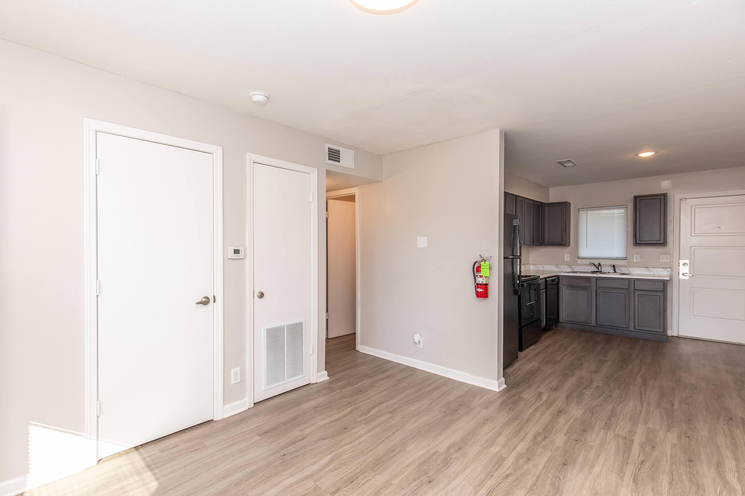 Interior view of a modern apartment showing a living area with light wood flooring, a kitchen area in the background, a white front door, and an open hallway. The kitchen features dark cabinets and stainless steel appliances. A fire extinguisher is mounted on the wall near the entrance.