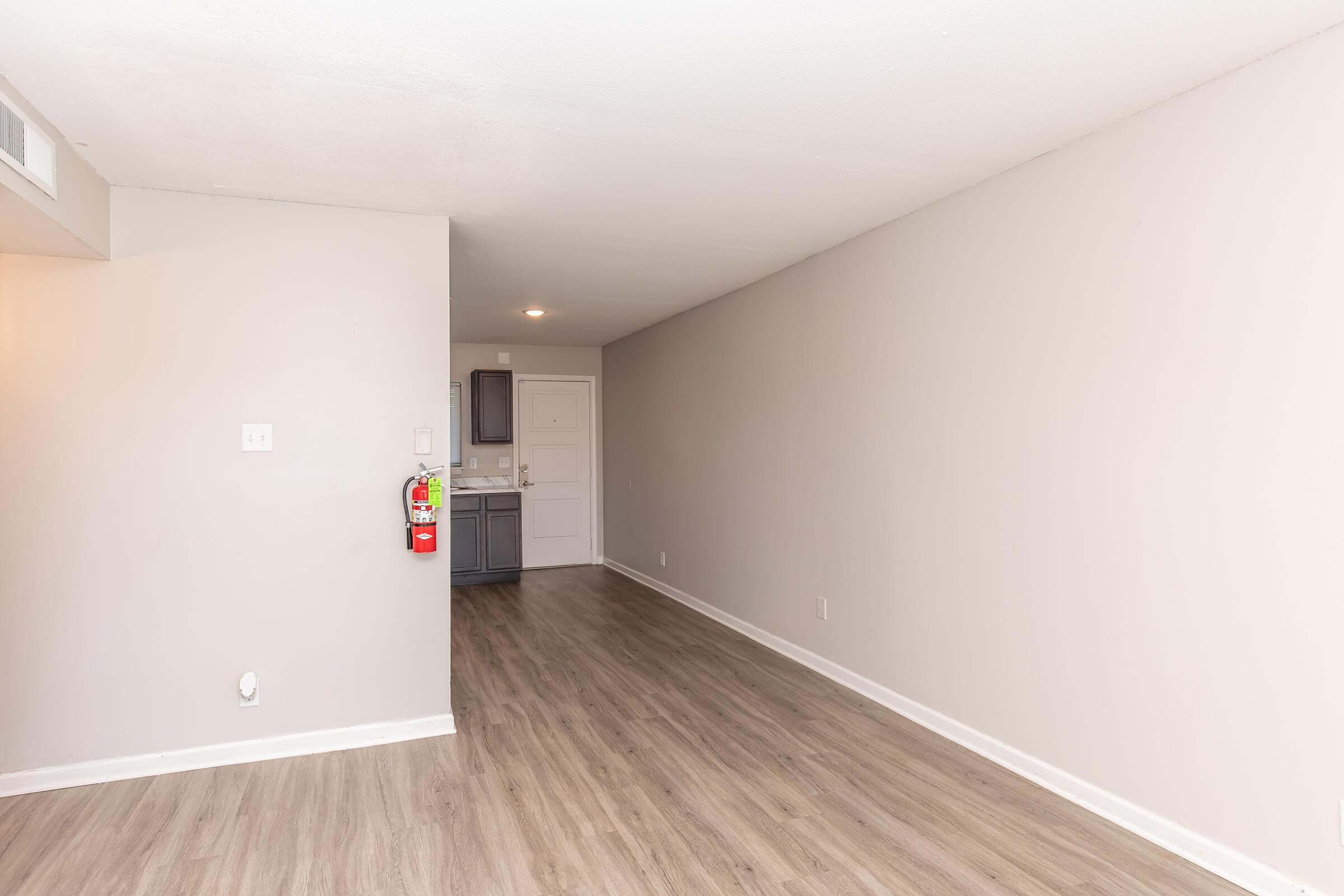 A spacious, empty room with light gray walls and laminate flooring, featuring a small kitchenette in the background. There is a fire extinguisher mounted on the wall near the entrance. The room is well-lit with a ceiling light, creating an inviting atmosphere.
