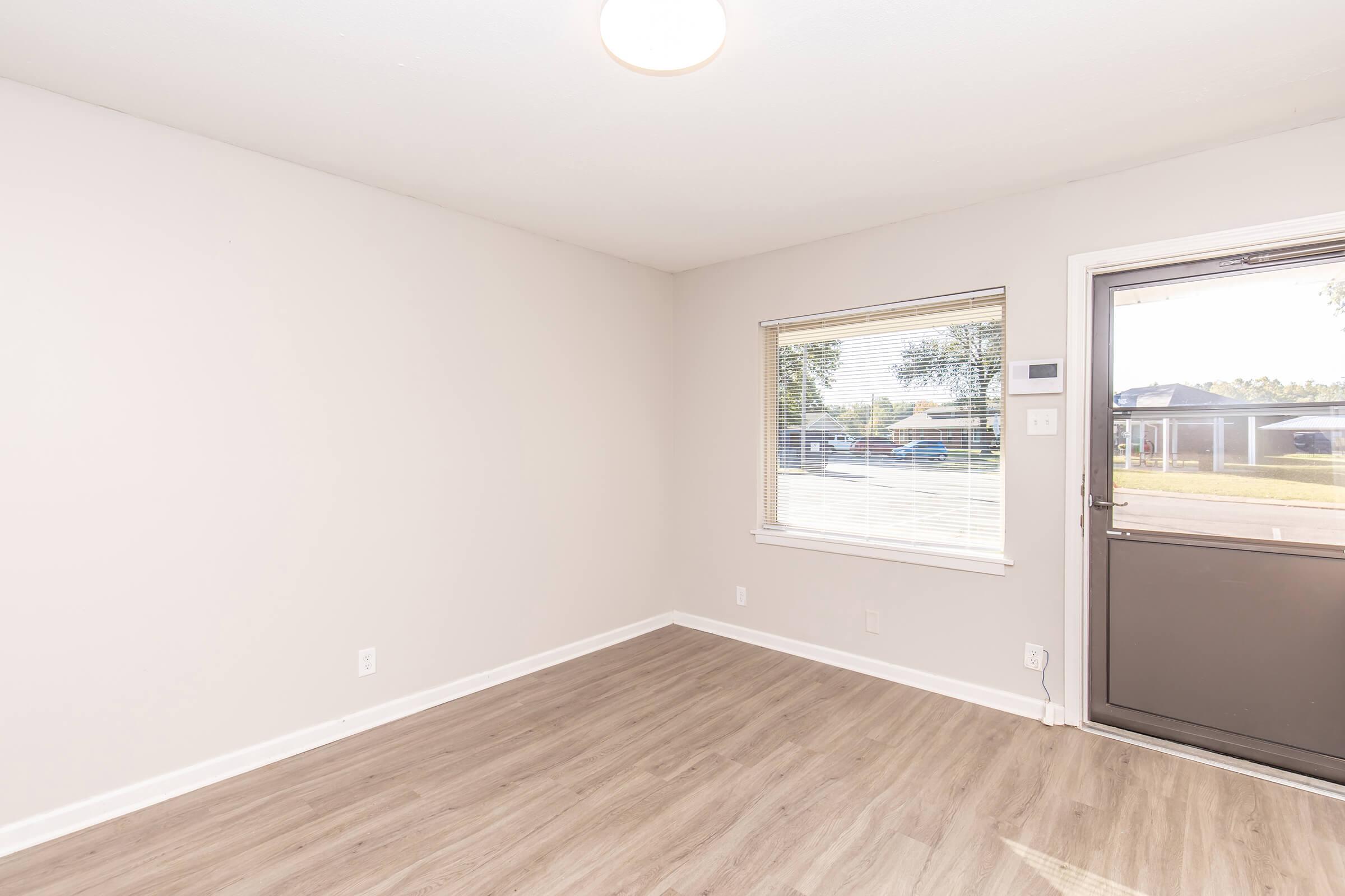 Empty room with light-colored walls and laminate flooring. A large window allows natural light to enter, with a view of the outdoors. The front door is visible, featuring a glass panel. The space is minimalistic, ideal for personalized decor.