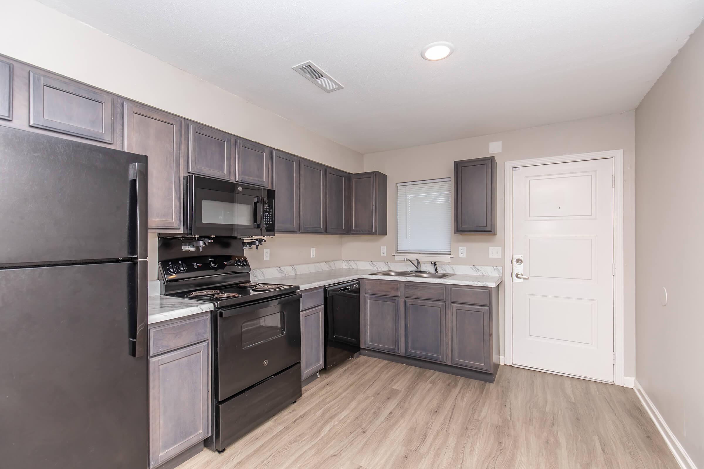 A modern kitchen featuring dark wooden cabinets and stainless steel appliances, including a black refrigerator, stove, and microwave. The countertop is light-colored, and there is a window above the sink. A door leading outside is visible on the right side of the image. The flooring is light wood laminate.