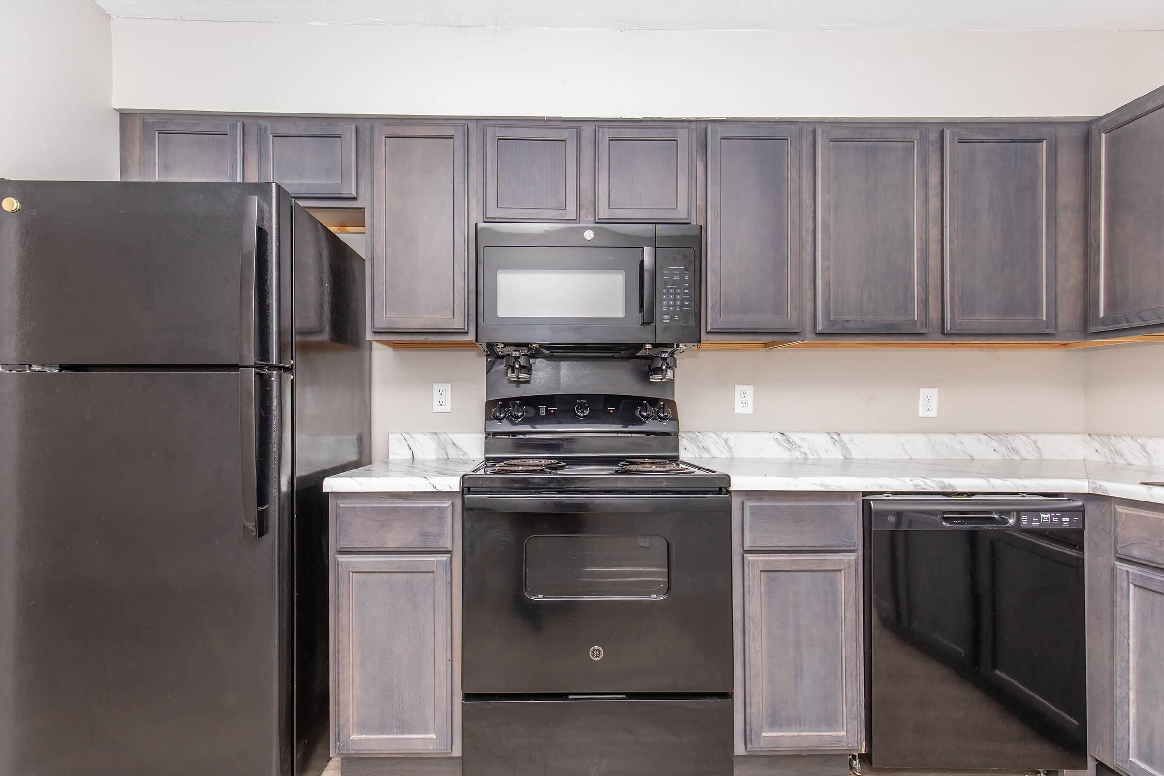Modern kitchen featuring black appliances including a refrigerator, microwave, oven, and dishwasher. The cabinetry is dark wood with a marble countertop. The layout is compact and functional, showcasing a clean and organized space.