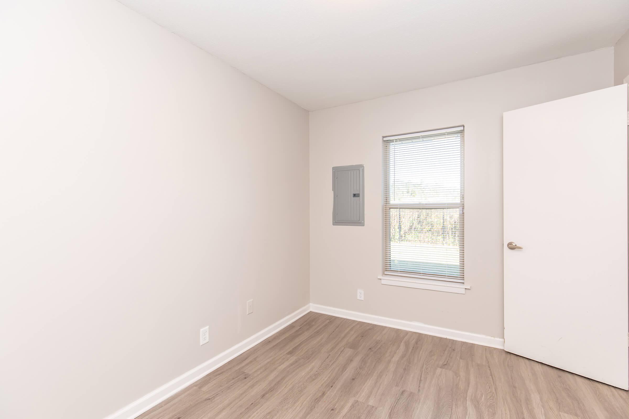 Empty room with light-colored walls, large window with blinds, and a white door. A small electrical panel is mounted on the wall near the window. The floor is covered with a light-colored laminate or hardwood. Natural light enters through the window, highlighting the room's simplicity.