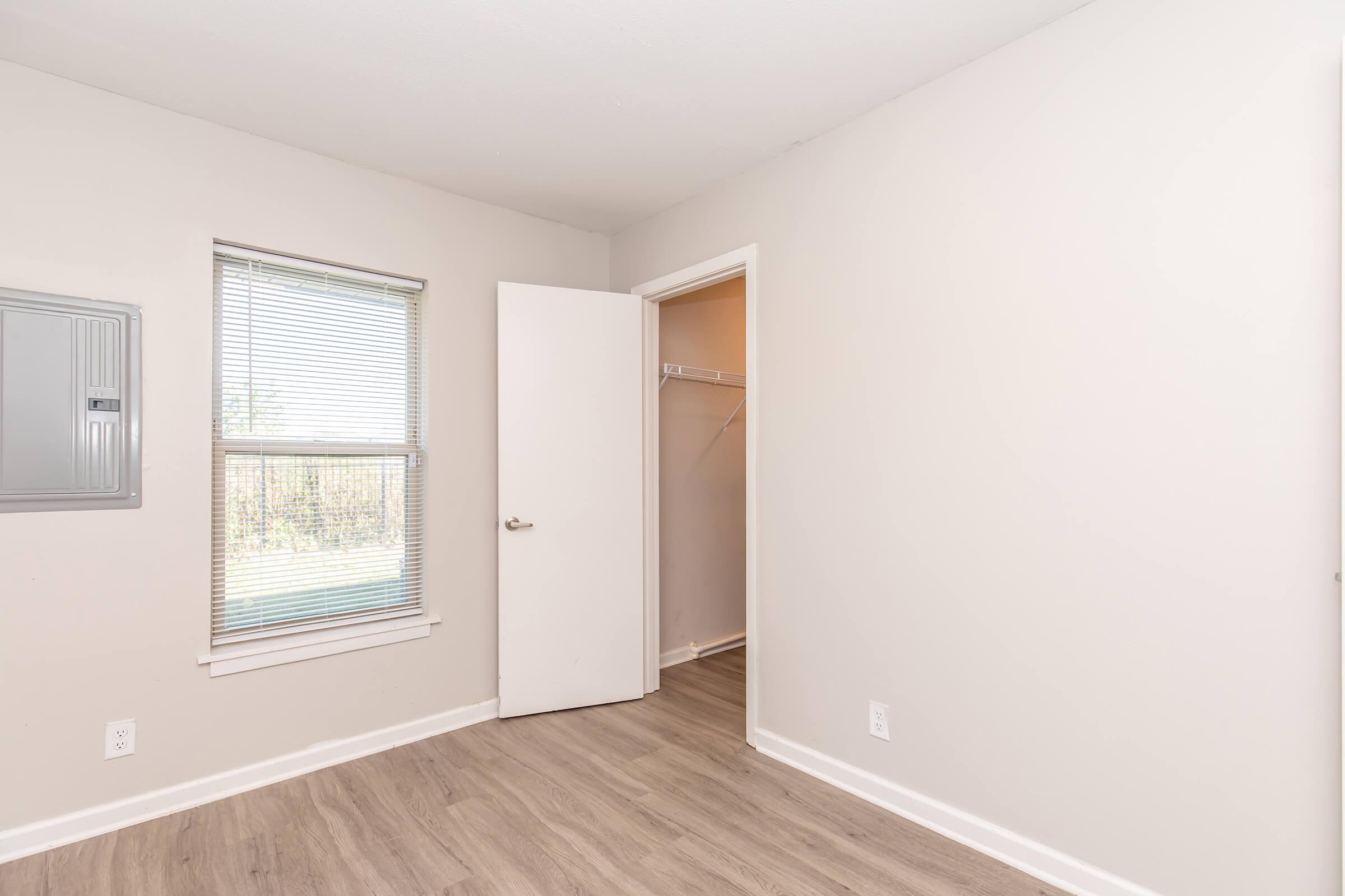 Bright, empty room with light-colored walls and a hardwood floor. A window with blinds provides natural light, and a door leads to a small closet. The room has minimal furnishings, creating a spacious feel.