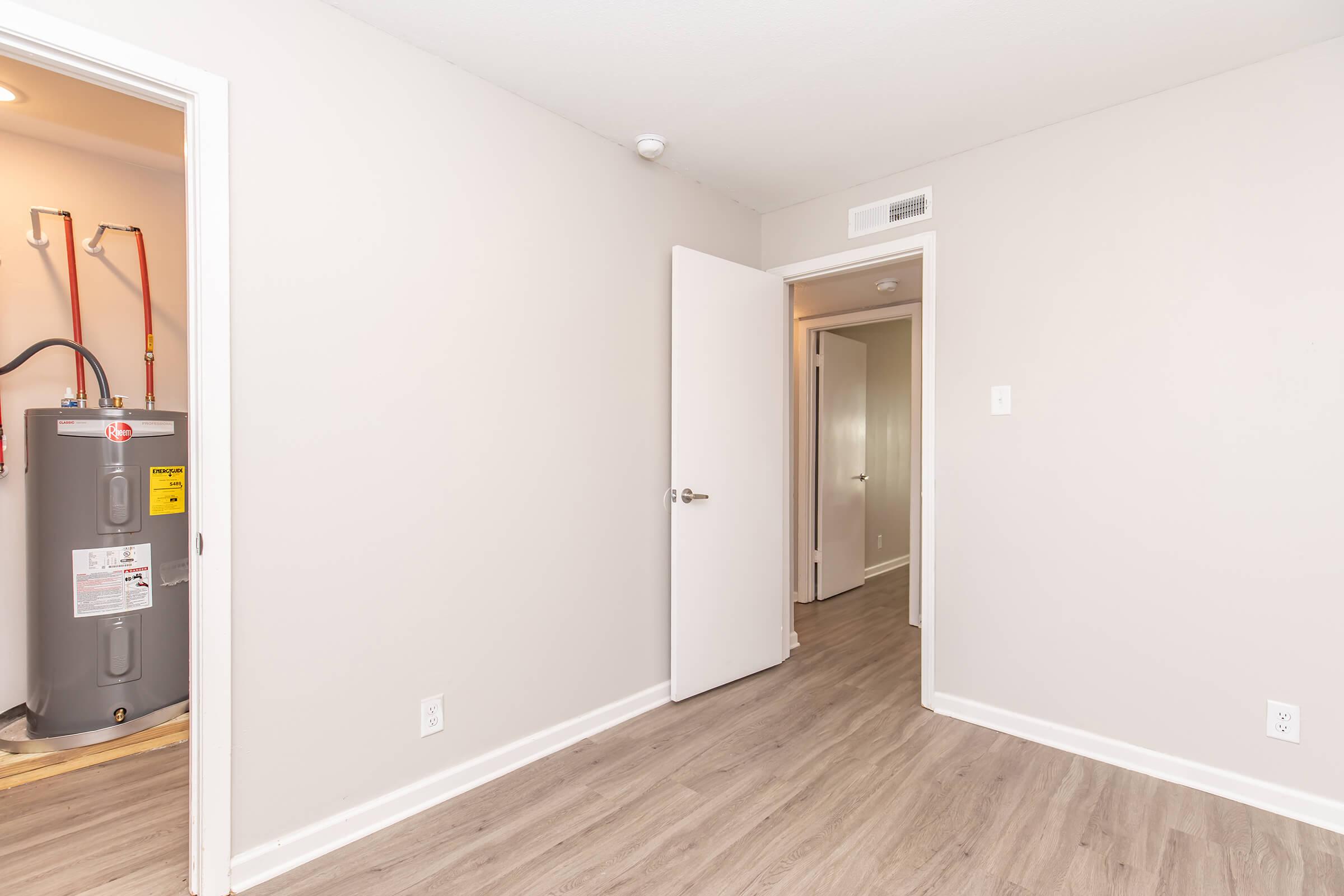 A clean, minimally decorated room with light gray walls and laminate flooring. A door is open to a hallway, while another door leads to a utility area with a water heater visible. Natural light brightens the space, contributing to a modern, inviting atmosphere.