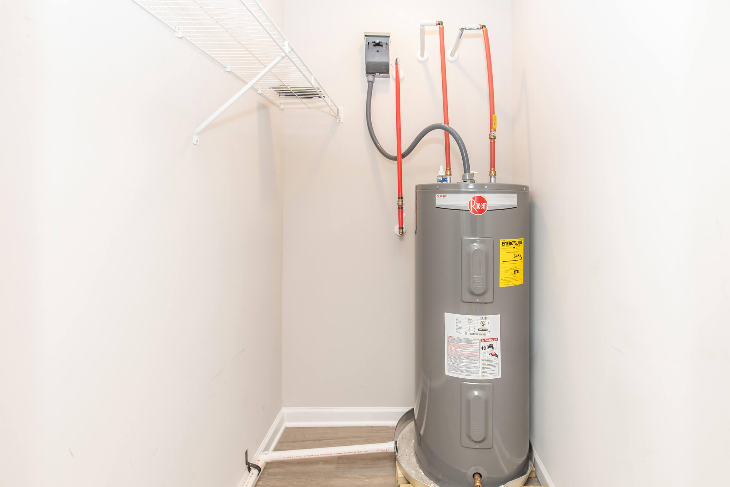 A gray water heater with visible pipes and a yellow label, situated in a small closet with light-colored walls and a wooden floor. A metal shelf is mounted on the wall beside the water heater.