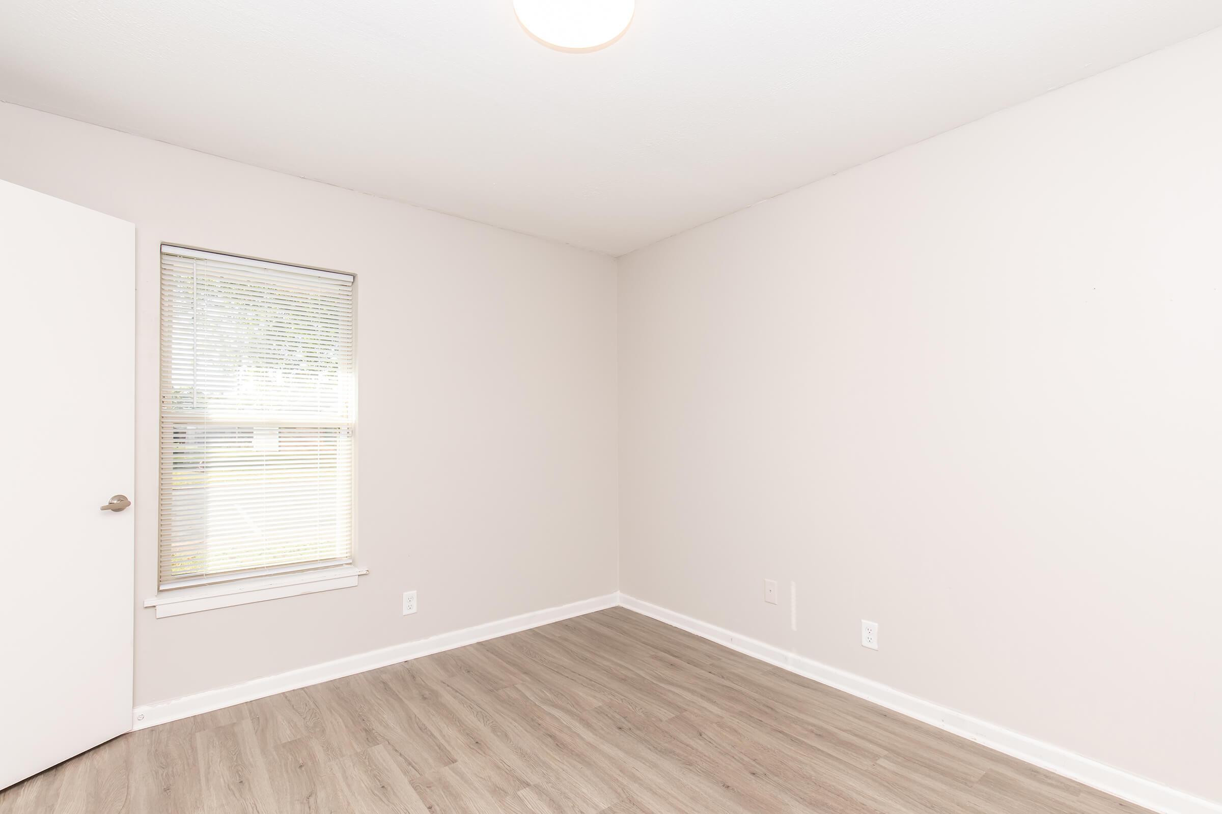 Empty room with light gray walls and a single window covered with blinds. The floor is light-colored laminate. A plain door is visible on the left side, and a ceiling light fixture is present. The space has a clean, minimalistic appearance with no furniture or decorations.