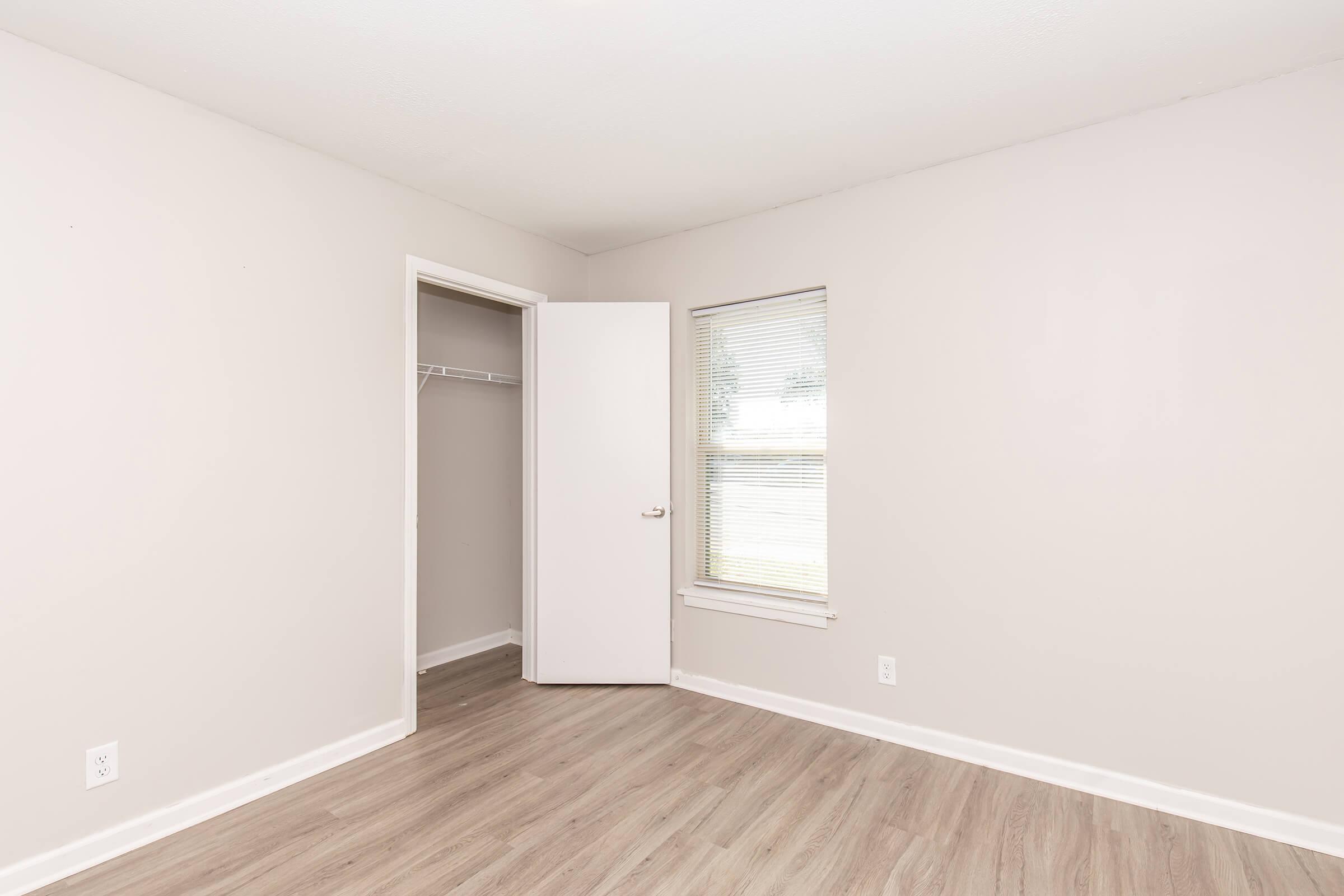 Empty room with light beige walls and wooden flooring. There is a white door slightly ajar leading to a closet and a window with blinds allowing natural light. The space is minimal and uncluttered, giving it a clean and open feel.