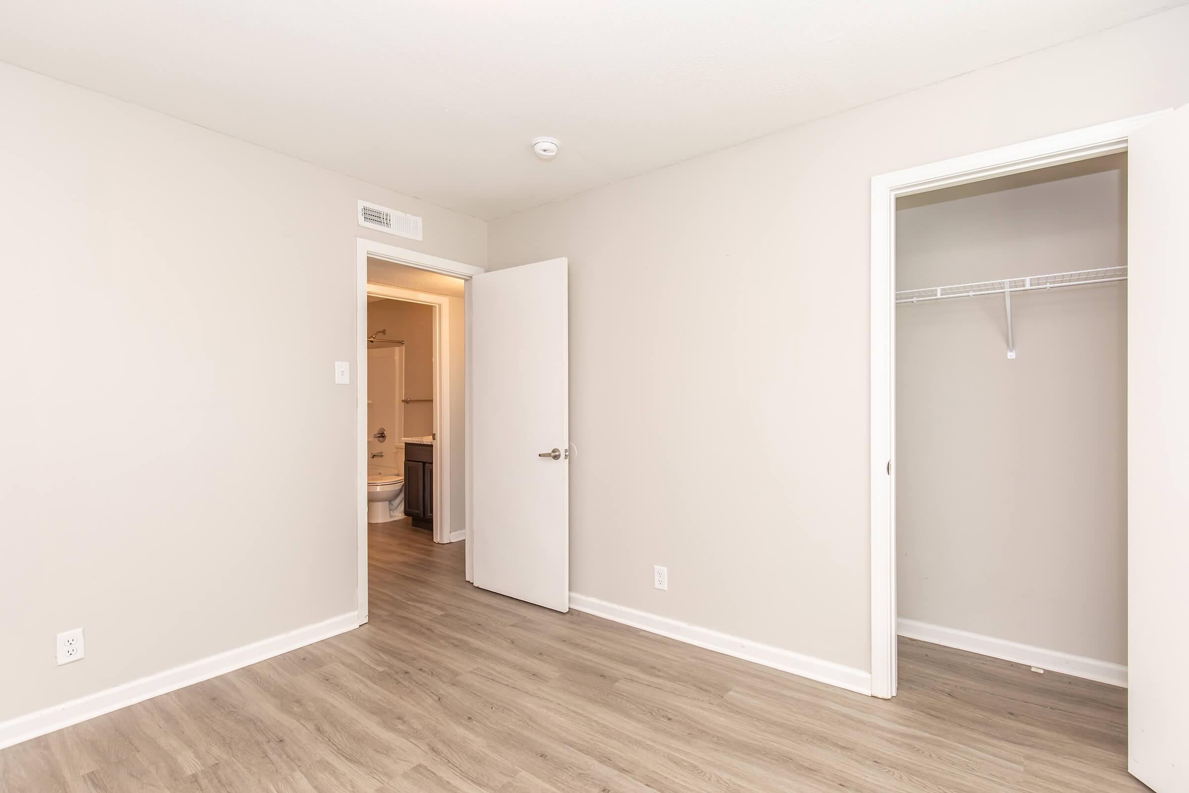 Empty room with light gray walls and wood-look flooring. A closed white door leads to a closet on the right, while an open door in the background reveals a bathroom. The room is well-lit and spacious, creating a minimalist and inviting atmosphere.