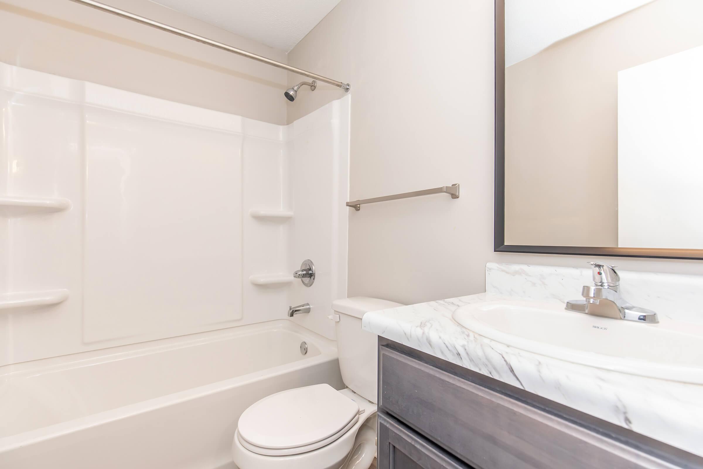 A clean and modern bathroom featuring a white bathtub, toilet, and dark wood vanity with a marble countertop. There is a wall-mounted mirror above the sink and a shower curtain rod. The walls are painted a light neutral color, contributing to a bright and airy atmosphere.