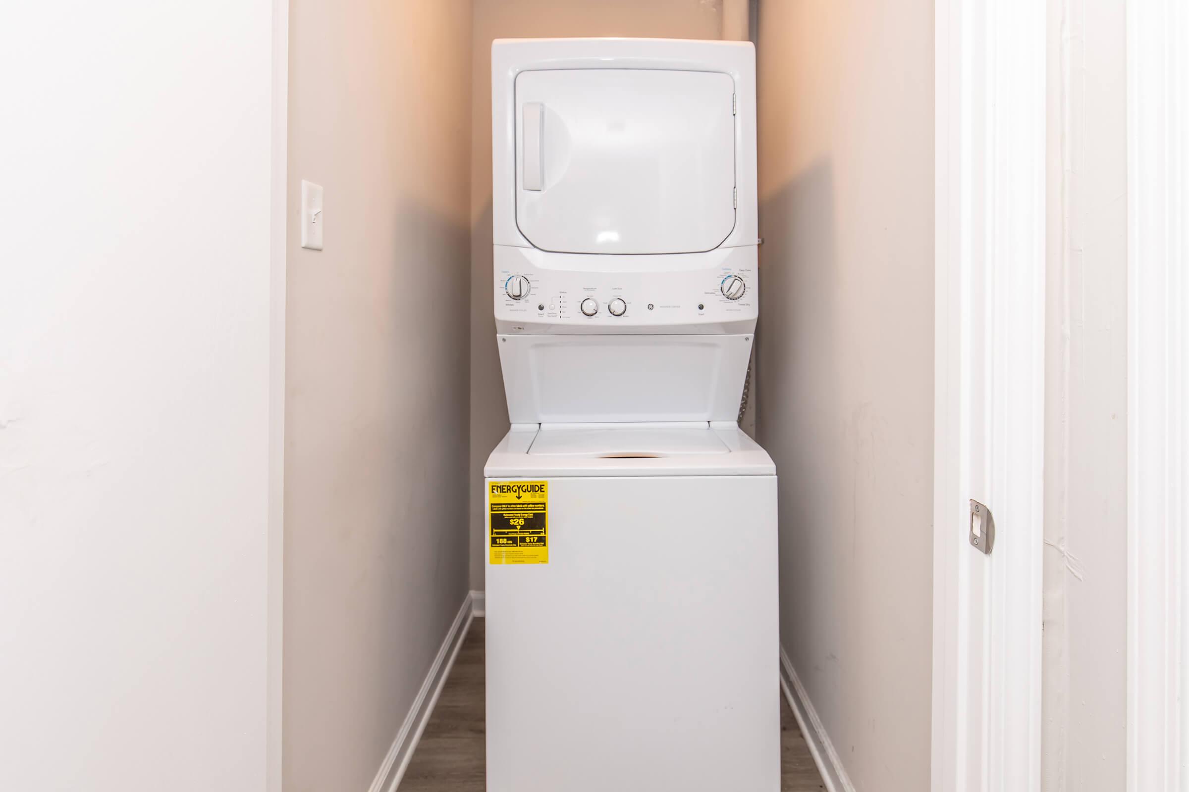 A stacked washer and dryer unit in a narrow hallway. The white appliances are positioned closely together, with light beige walls on either side. There is a safety warning sticker on the dryer. The flooring is a light wood-like material, adding to the modern aesthetic of the space.