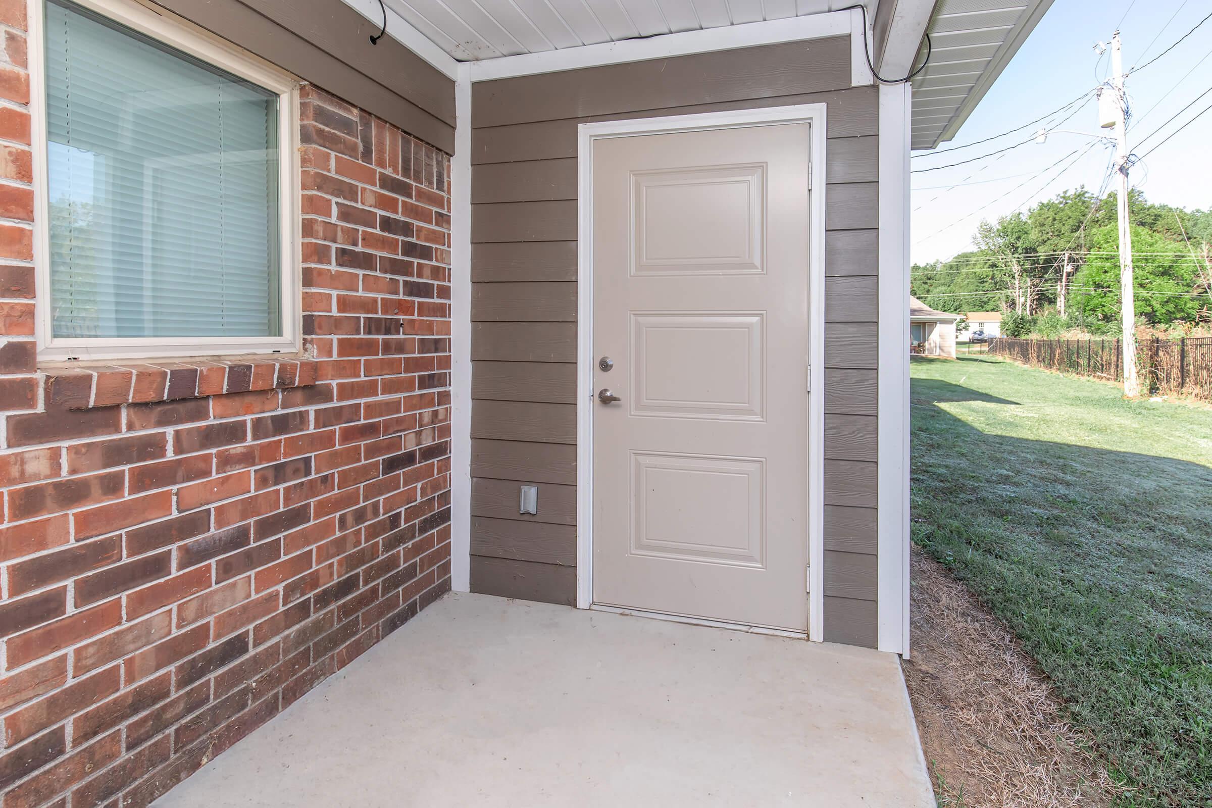 Exterior view of a small entryway featuring a beige door with a single doorknob, surrounded by a brick wall on one side and horizontal siding on the other. The porch is made of concrete, with grass visible on the side. Power lines and trees can be seen in the background under a clear sky.
