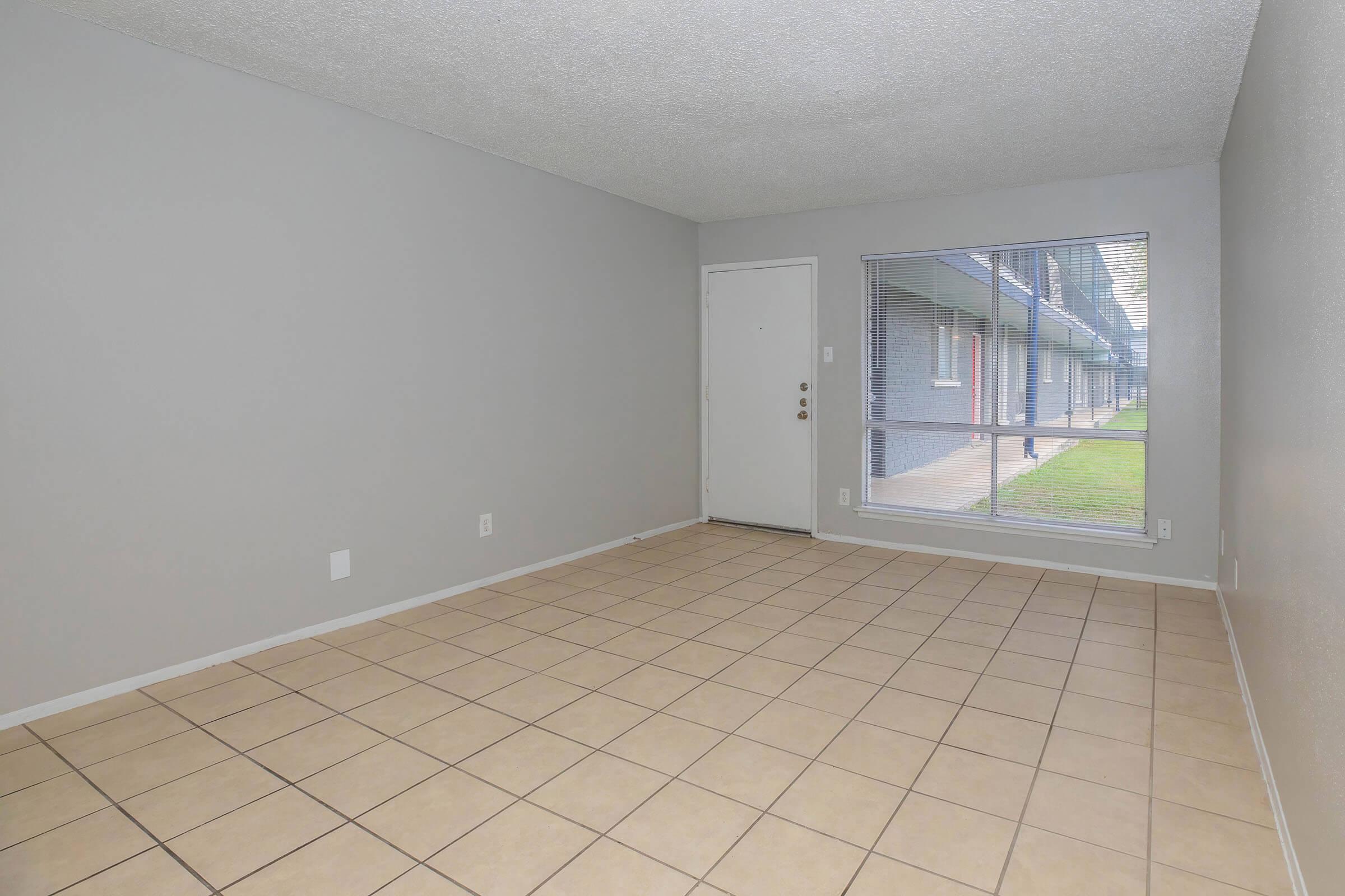 Empty room with gray walls and tile flooring. A door is visible on the left side, and a large window on the right lets in natural light, showcasing an exterior view of grass and a building facade. The space is clean and unfurnished, giving it a spacious feel.
